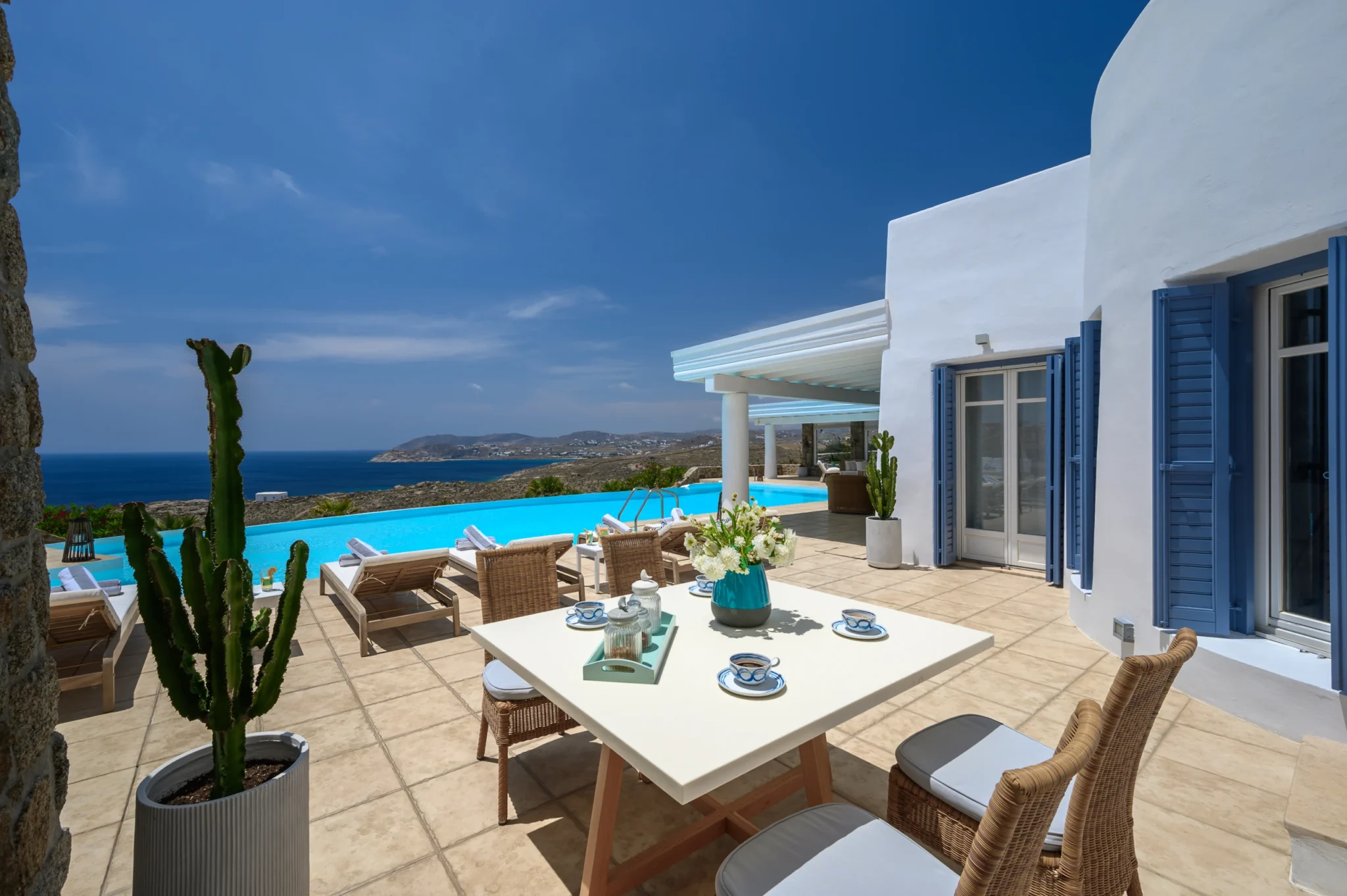 Outdoor table set with blue and white cups beside open blue shutters, potted cacti and the pool terrace at Villa Mustique in Mykonos.