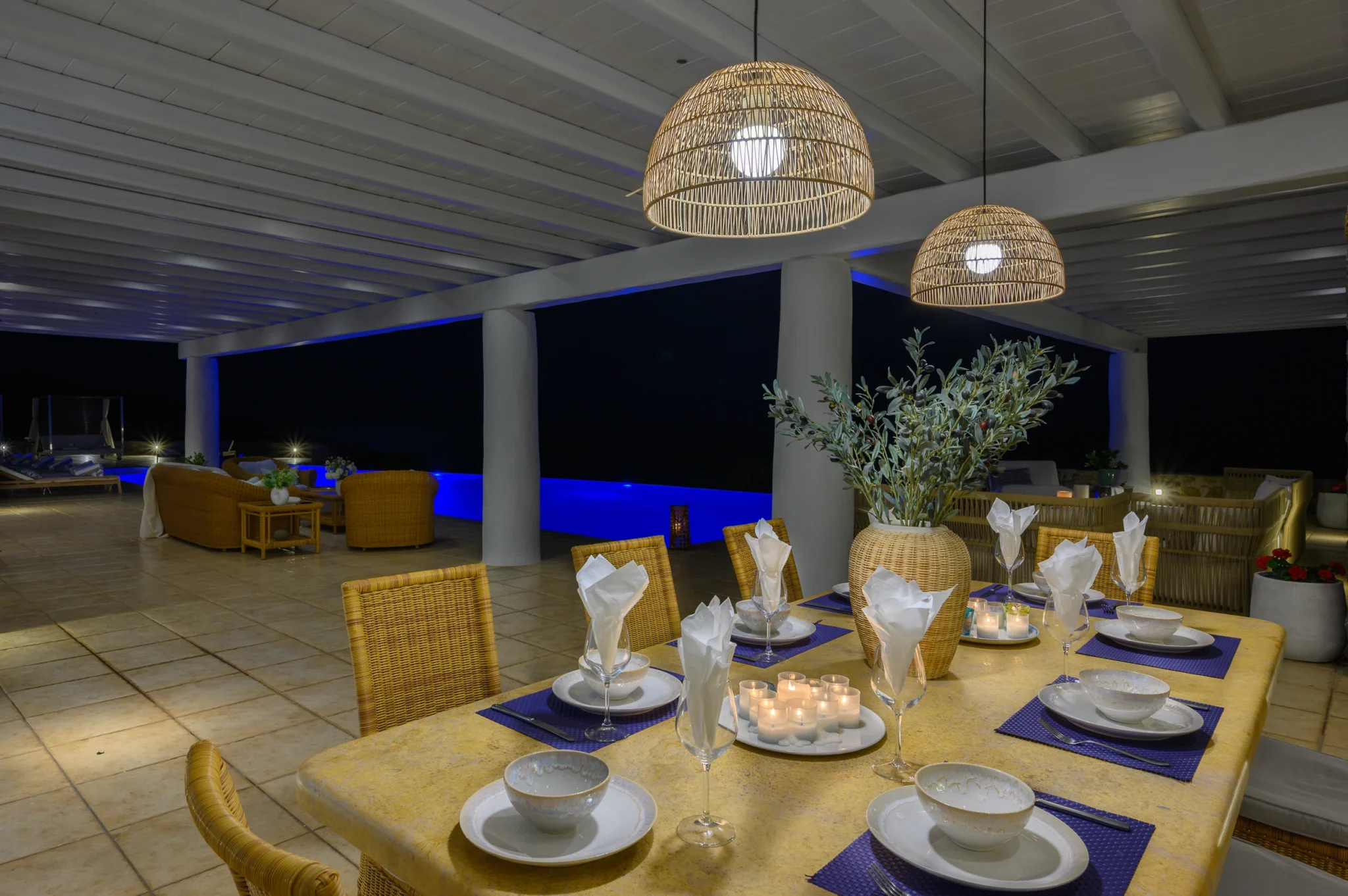Candlelit dining table with blue placemats and folded white napkins beneath woven pendant lights beside the illuminated pool at Villa Mustique in Mykonos.