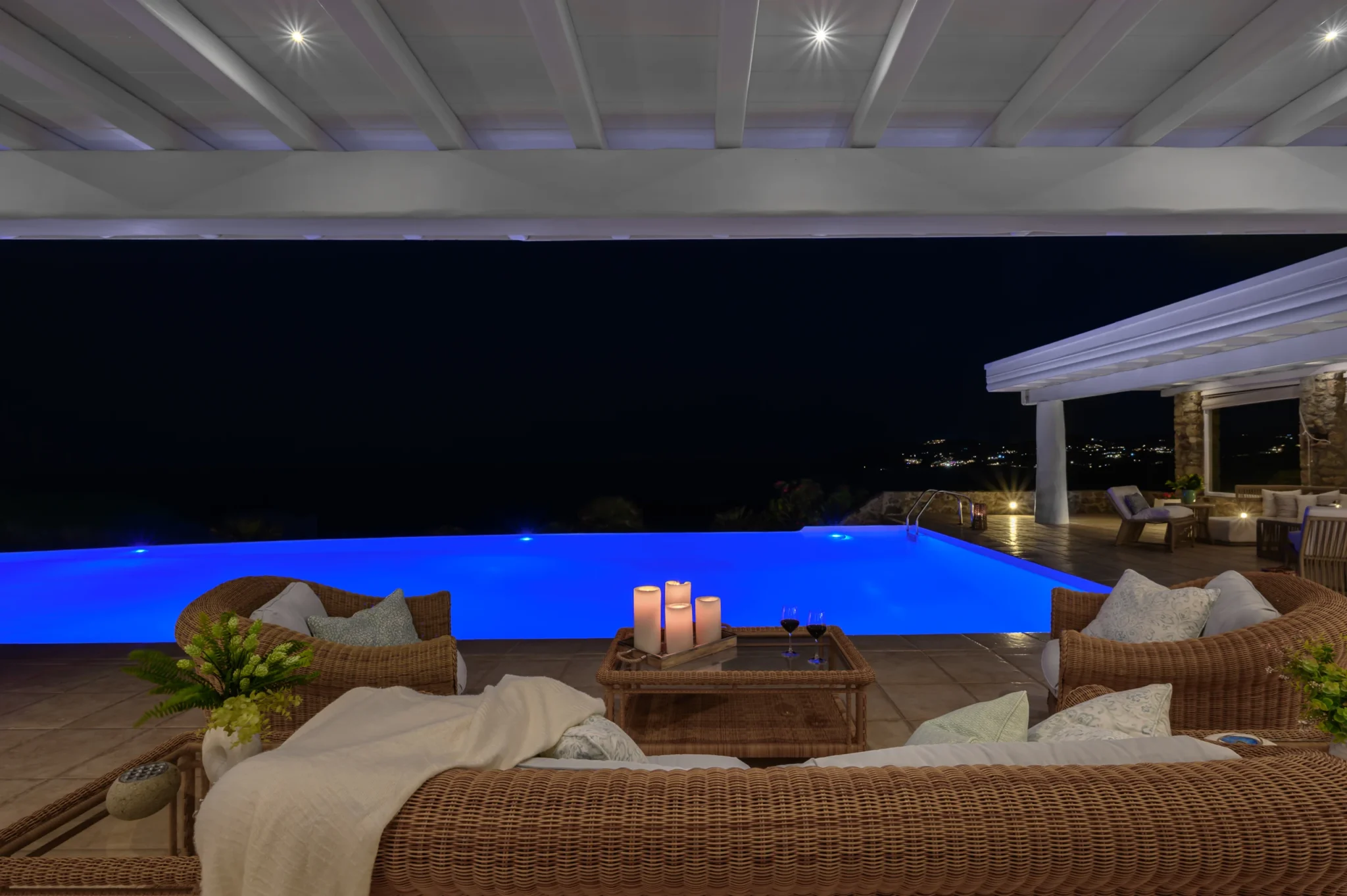 Wicker lounge beneath a white pergola at night, with candles and wine glasses beside the illuminated blue pool at Villa Mustique in Mykonos.