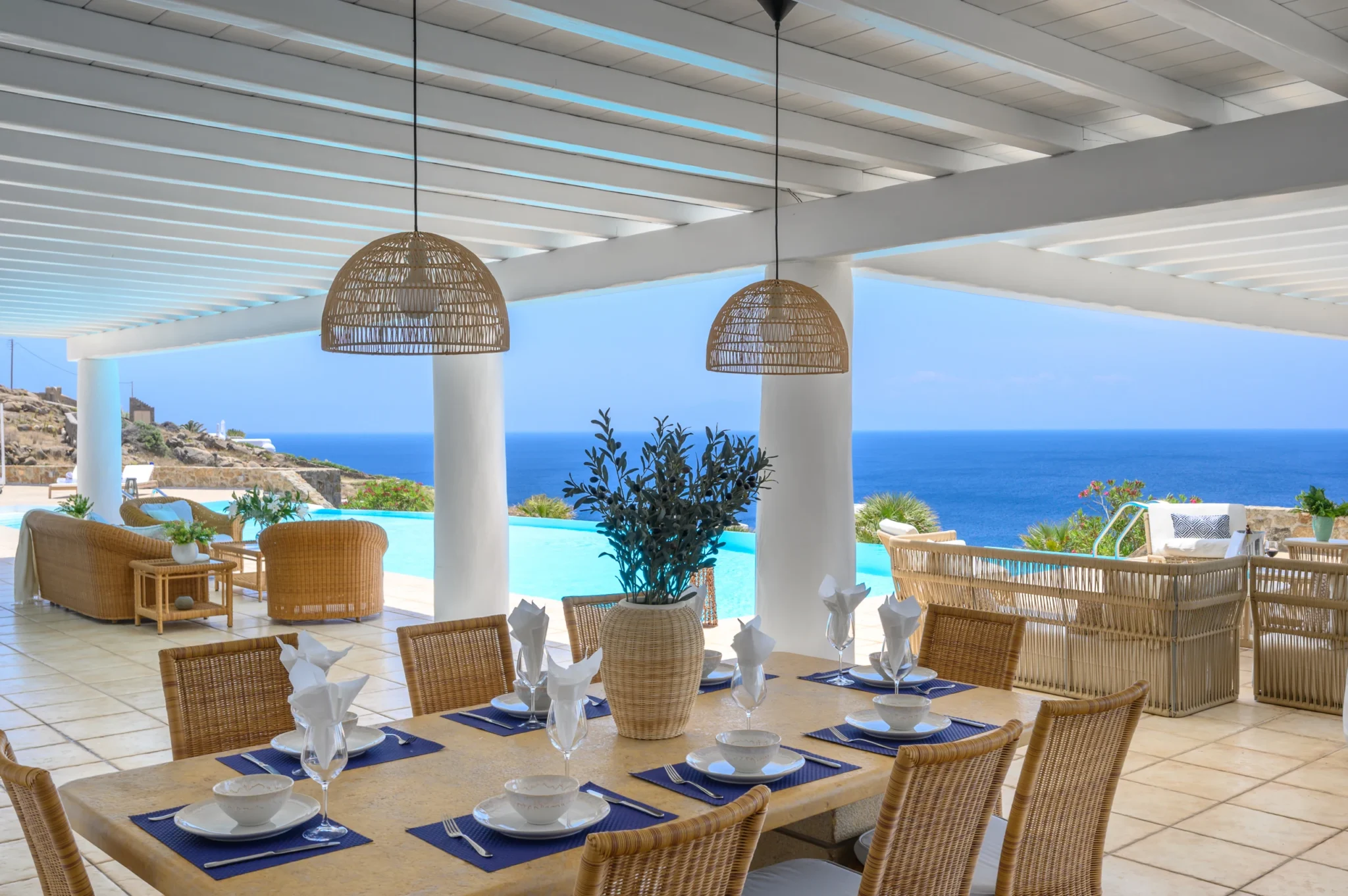 Set dining table beneath white pergola beams and woven pendant lights, overlooking the pool and sea at Villa Mustique in Mykonos.