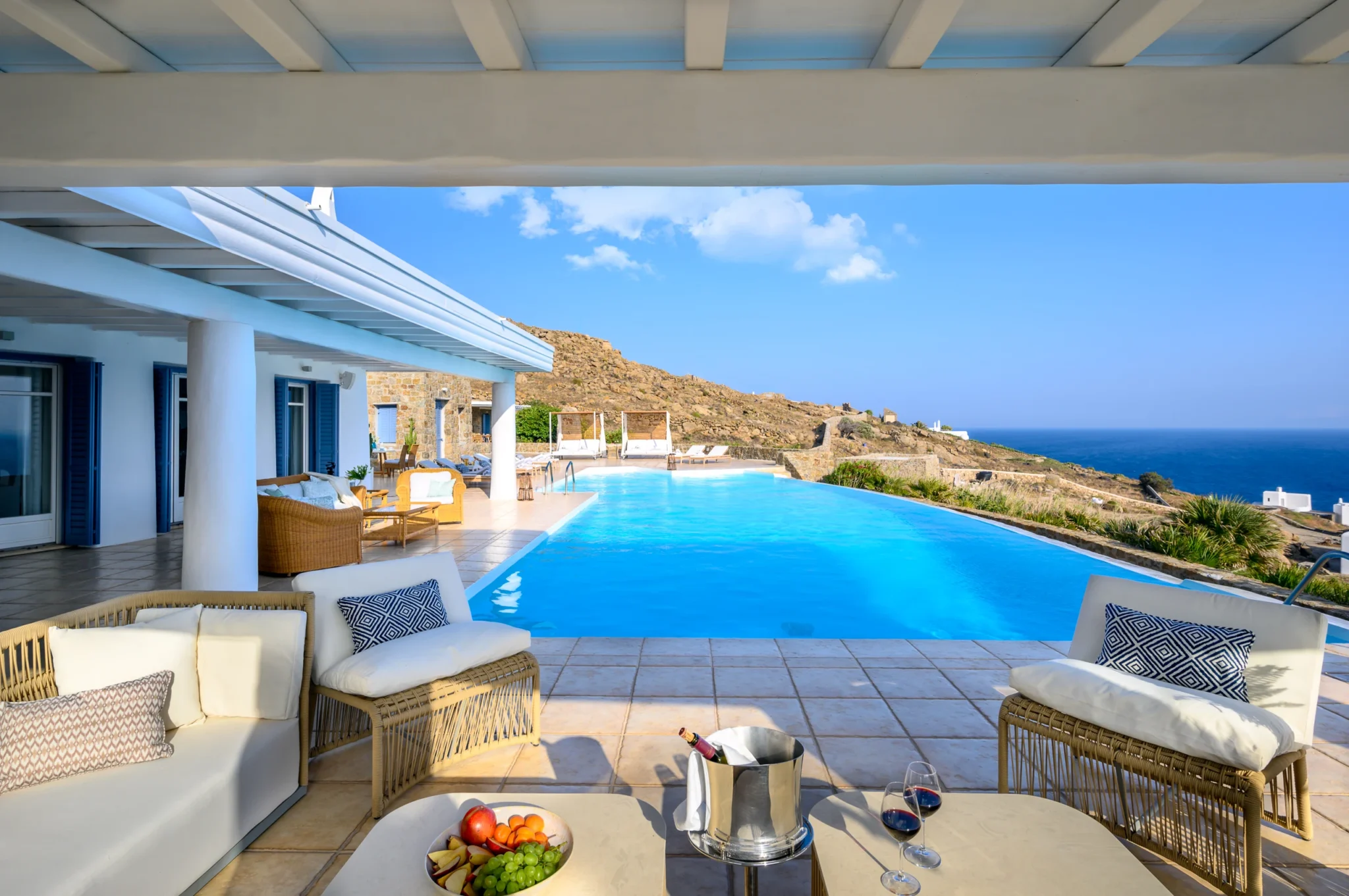 Covered woven lounge furniture with wine and fruit on the table, looking along the pool towards daybeds and the sea at Villa Mustique in Mykonos.