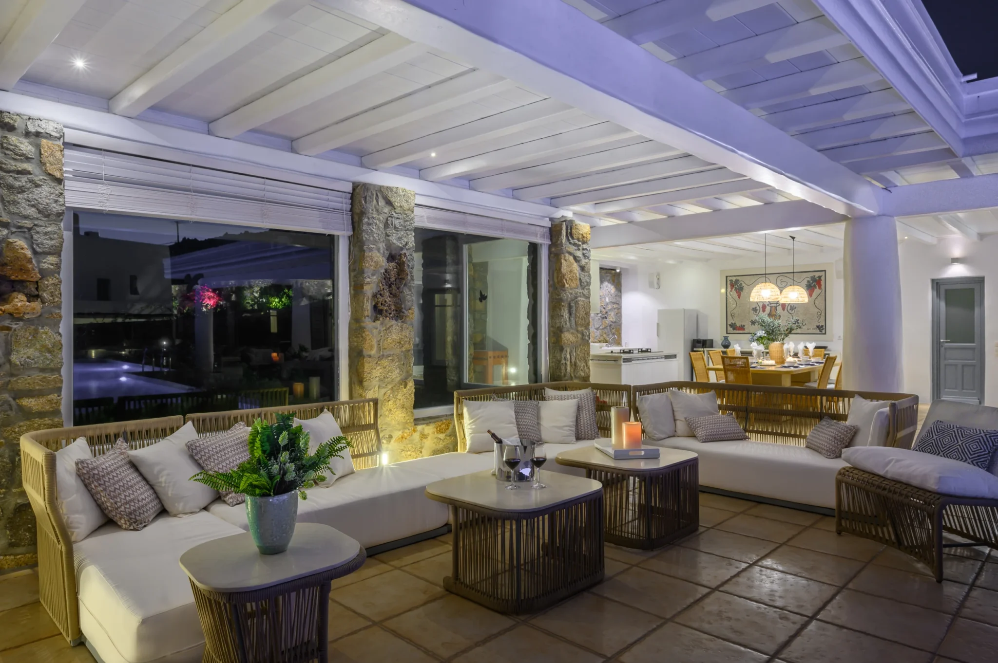 Woven lounge sofas beside stone columns and glazed screens, with candlelit tables and an illuminated dining area beyond at Villa Mustique in Mykonos.