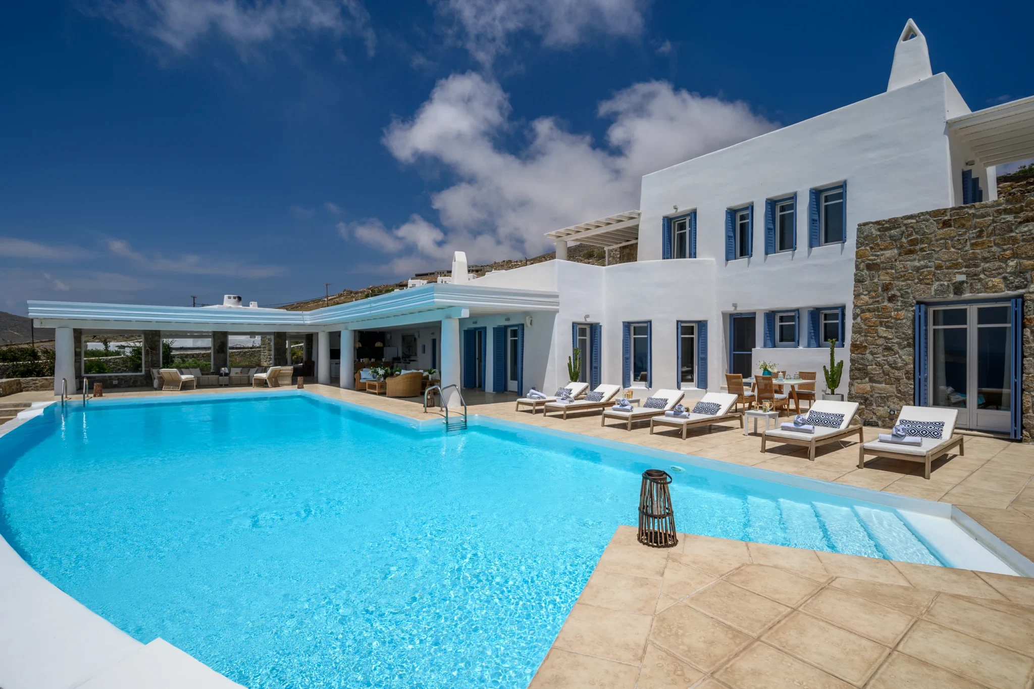 White and natural stone villa with blue shutters, timber sun loungers and a turquoise swimming pool at Villa Mustique in Mykonos.