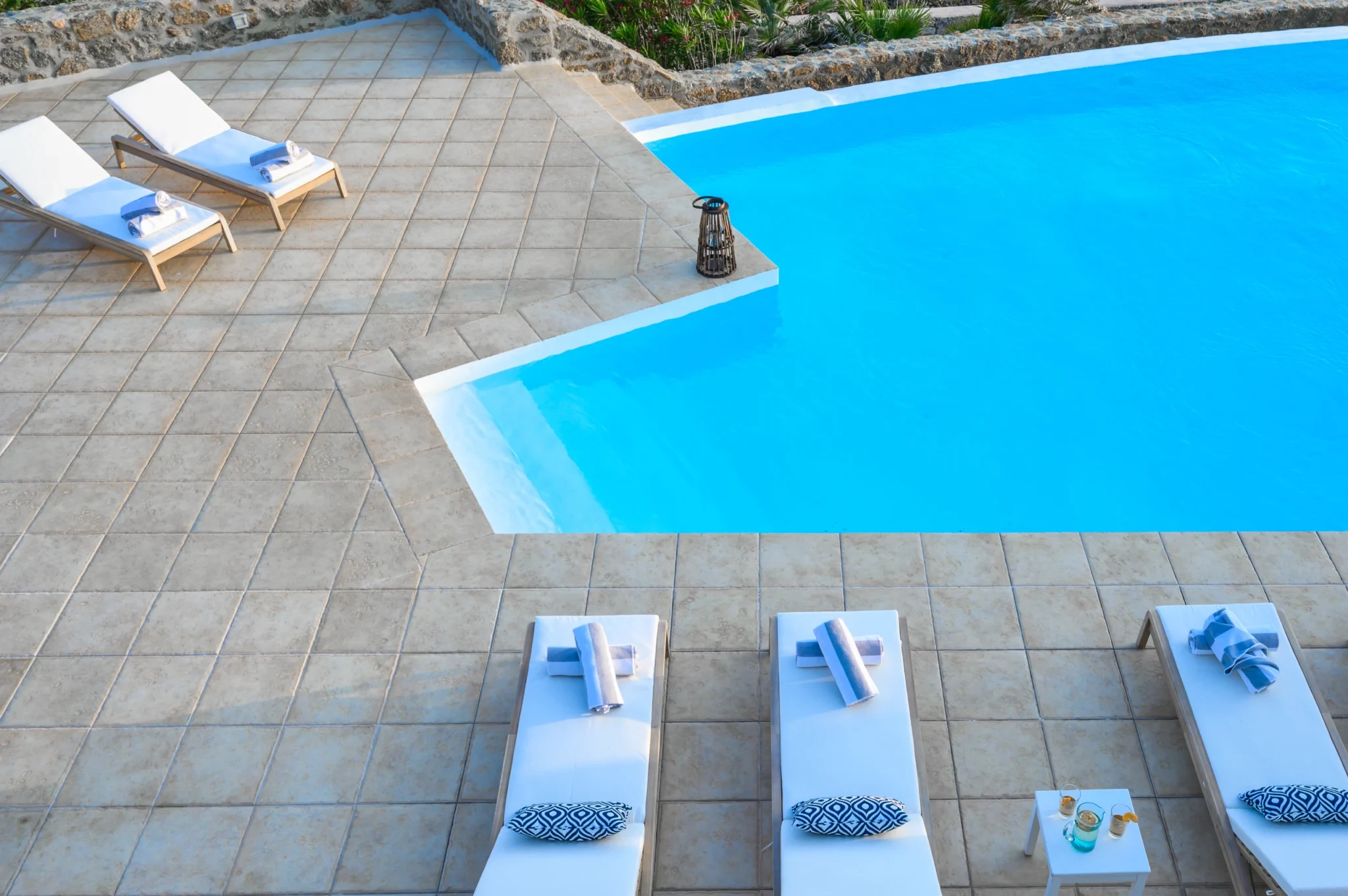 Elevated view of the angular swimming pool edge, tiled terrace and white-cushioned sun loungers at Villa Mustique in Mykonos.