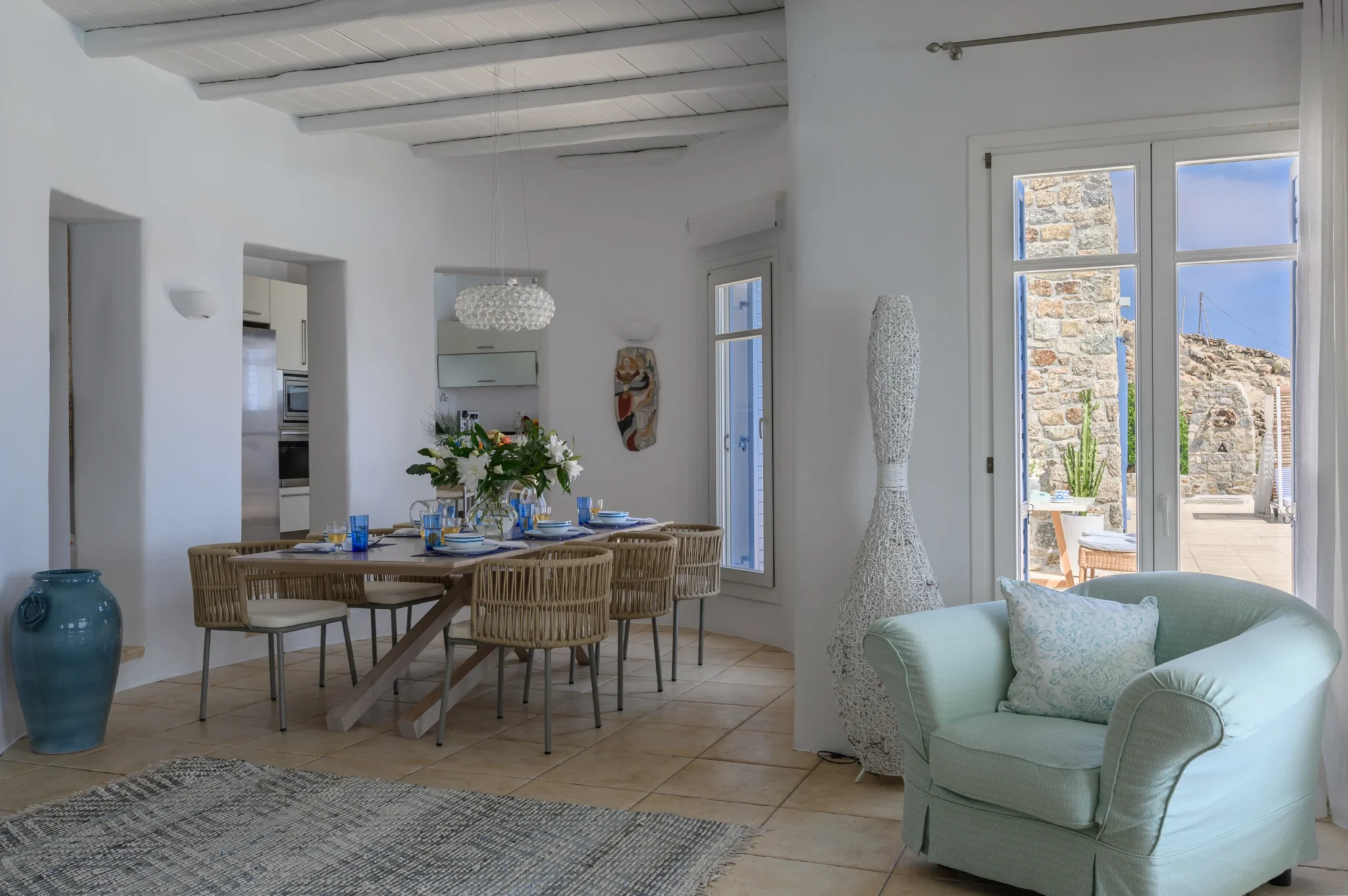 Interior dining table with woven chairs, blue glassware and white flowers, beside a glazed door to the terrace at Villa Mustique in Mykonos.