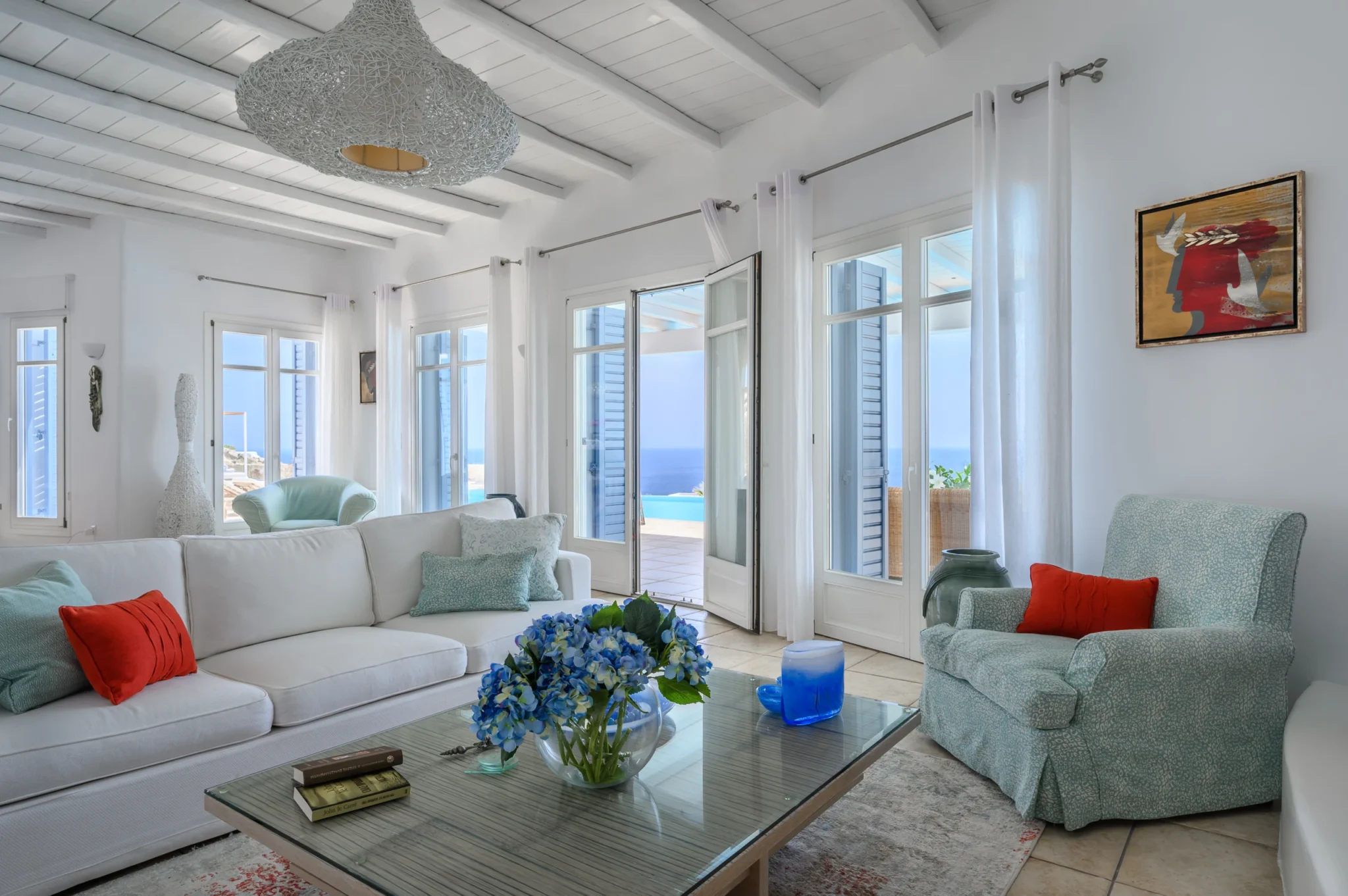 White living room with a sofa, pale turquoise armchair, colourful cushions and open doors towards the pool and sea at Villa Mustique in Mykonos.