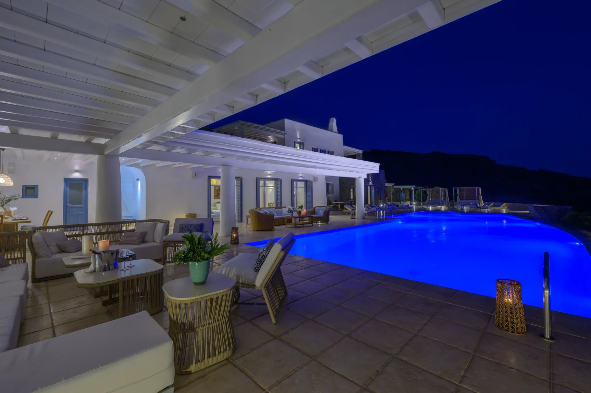 Covered lounge beneath white beams and columns, with the illuminated pool and villa doors at twilight at Villa Mustique in Mykonos.