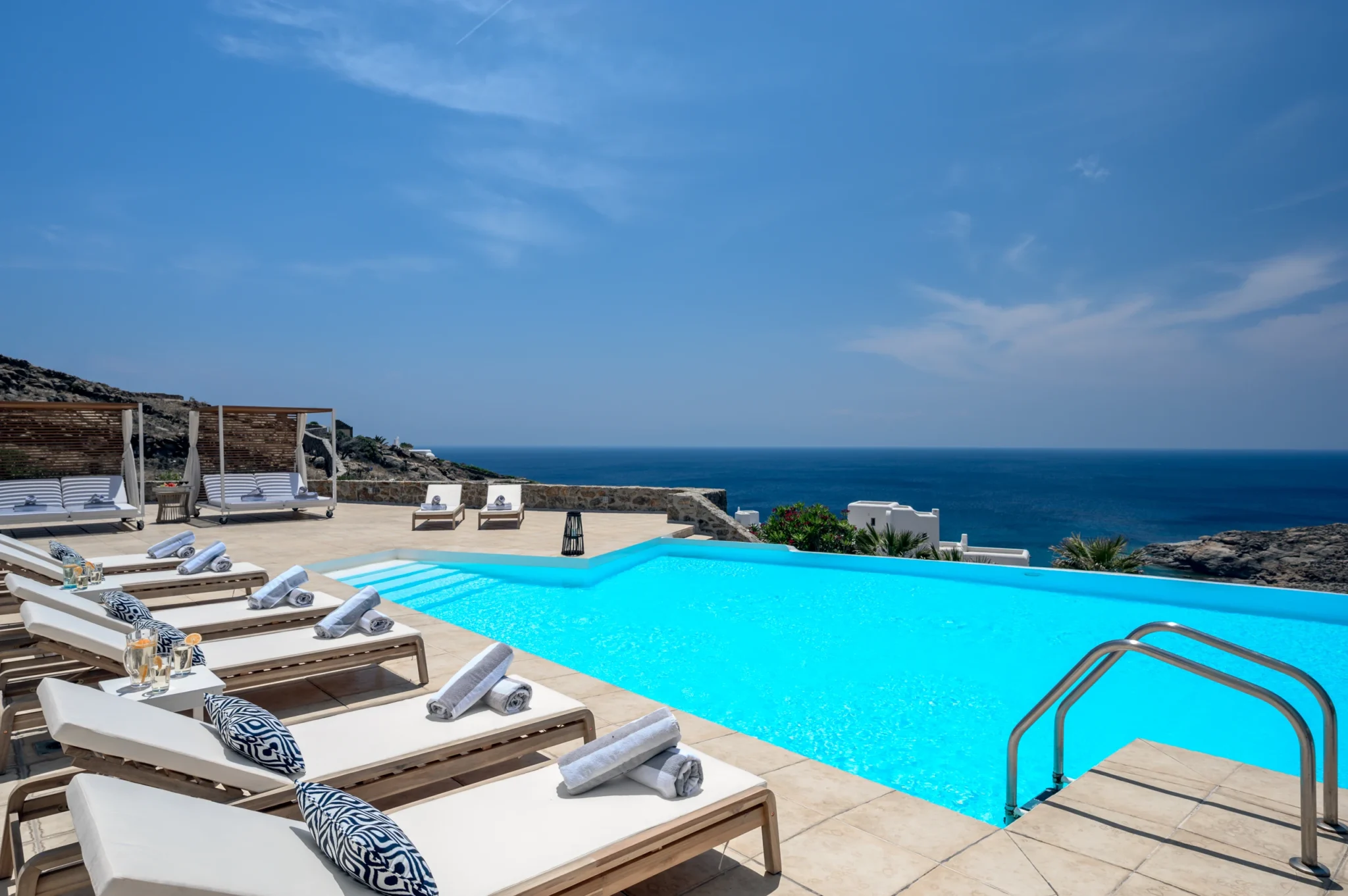 White-cushioned timber loungers, rolled towels and canopy daybeds beside the swimming pool overlooking the sea at Villa Mustique in Mykonos.