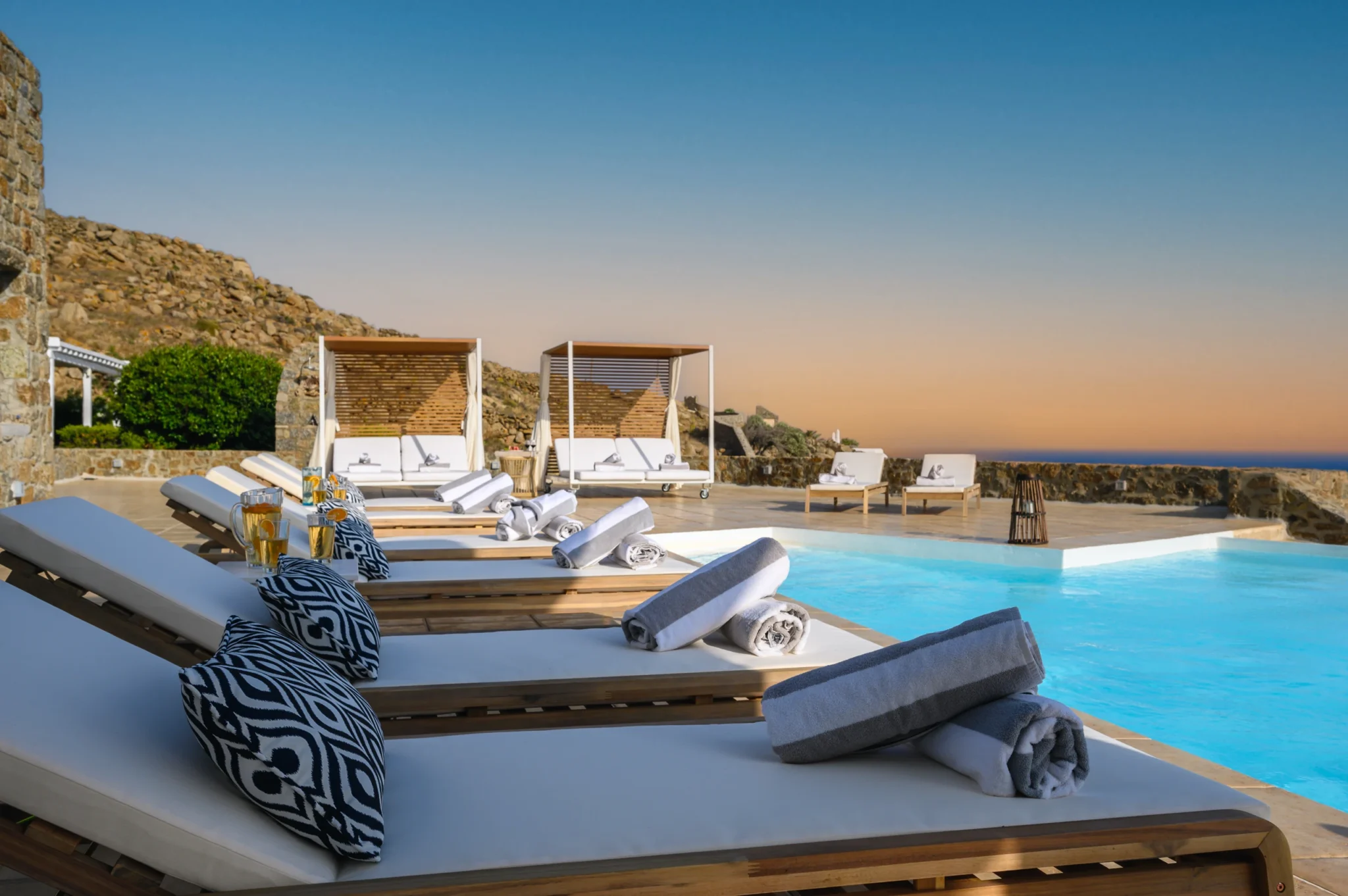 Timber loungers with white cushions and rolled towels beside the pool beneath a warm sunset sky at Villa Mustique in Mykonos.