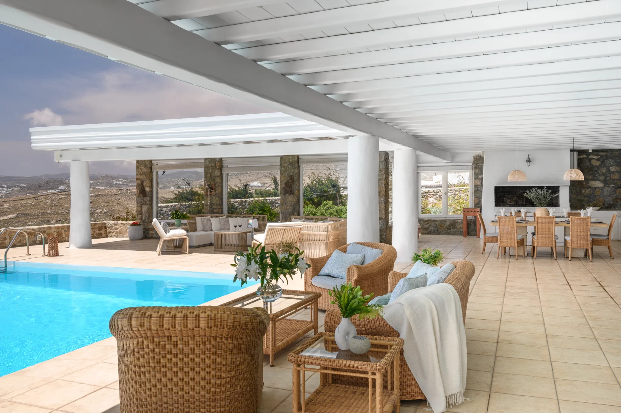 Wicker lounge furniture beside the swimming pool, with a covered seating area and outdoor dining table beyond at Villa Mustique in Mykonos.