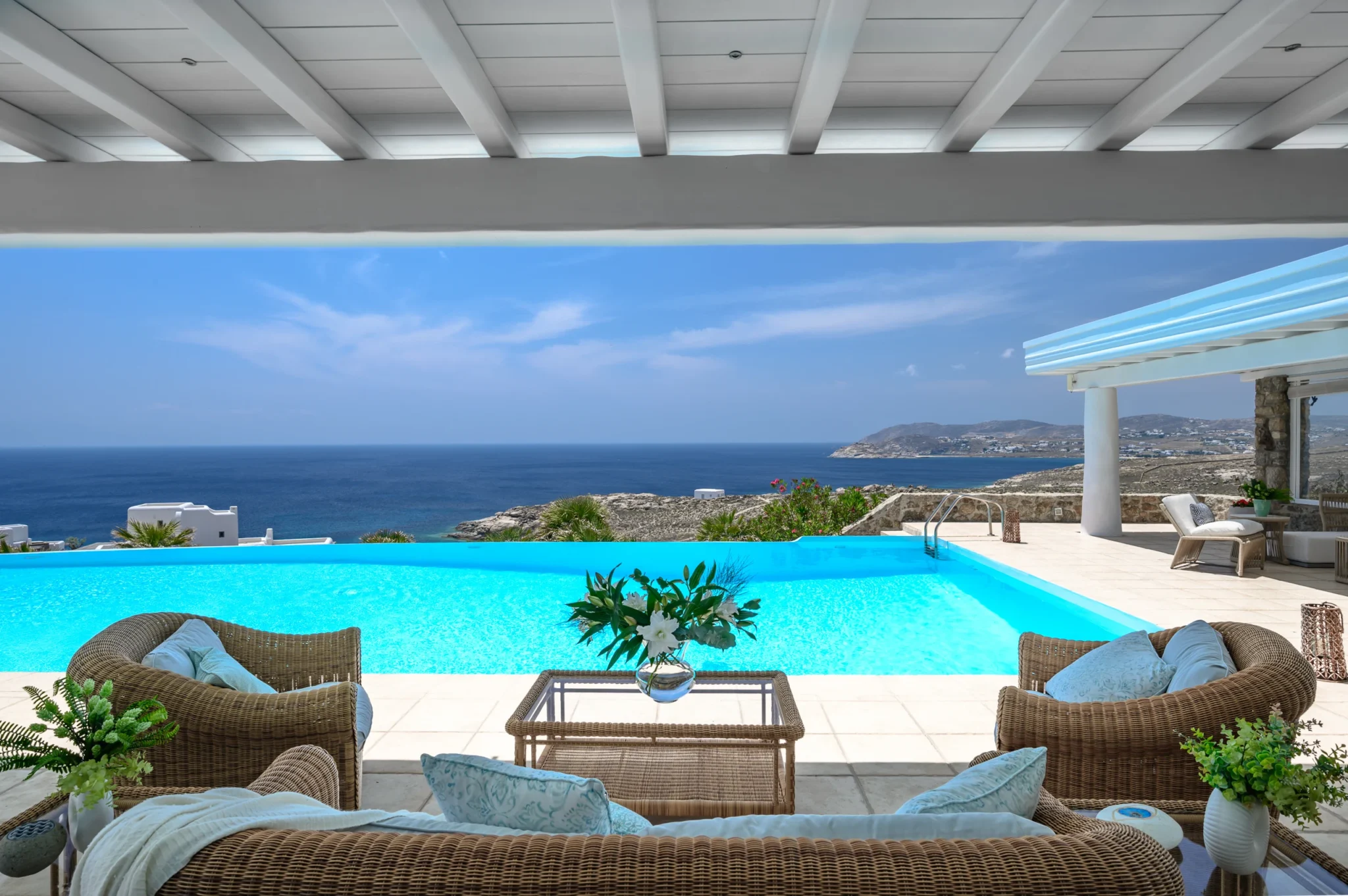 Wicker sofa and armchairs beneath a white pergola, facing the turquoise swimming pool and sea at Villa Mustique in Mykonos.