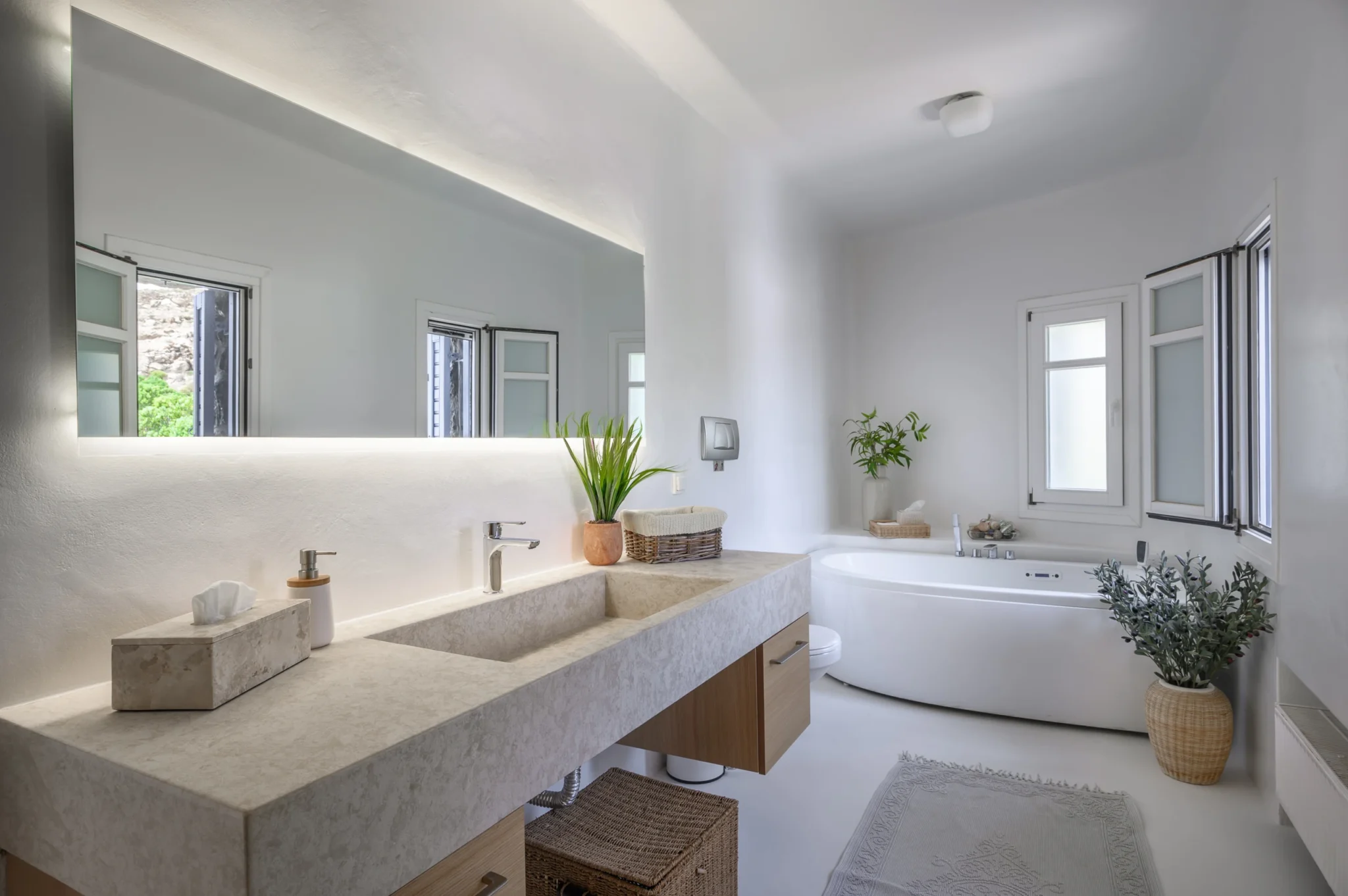 Stone bathroom vanity beneath a wide illuminated mirror, with a freestanding bathtub, white walls and natural baskets at Villa Mustique in Mykonos.