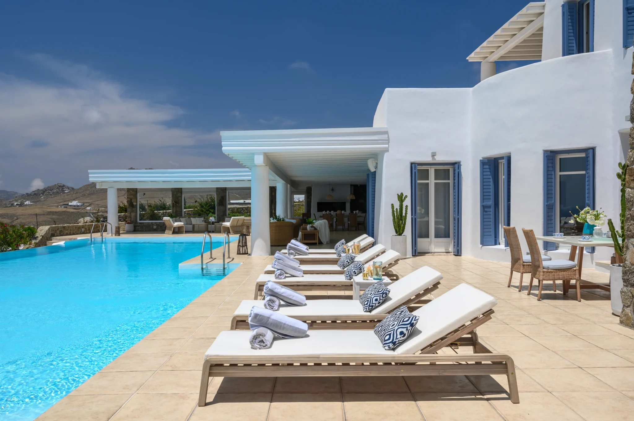 Row of timber sun loungers beside the pool, with blue-shuttered doors and white pergolas along the villa facade at Villa Mustique in Mykonos.
