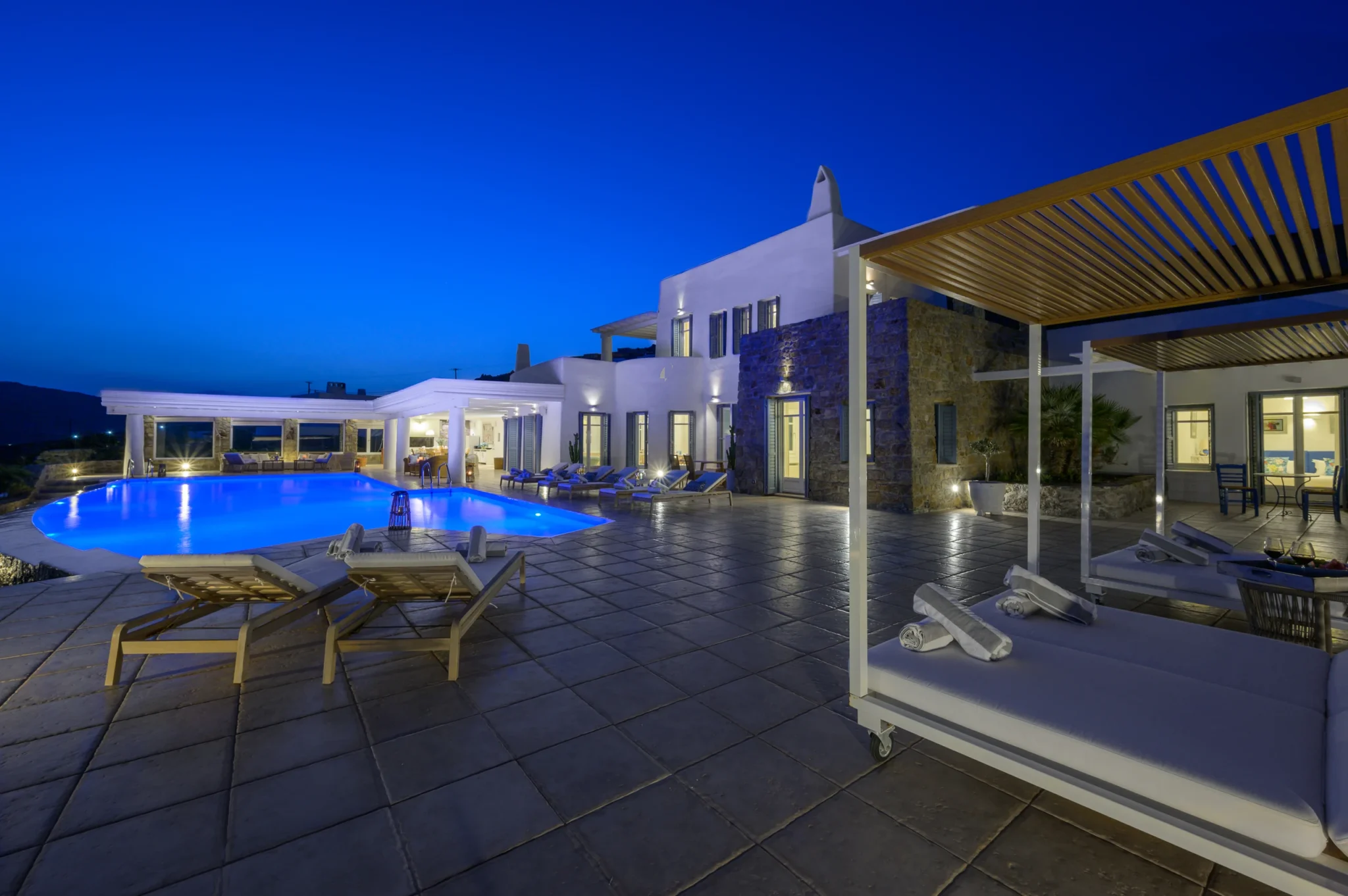 Illuminated blue swimming pool and white and stone villa, with canopy daybeds beneath a deep blue evening sky at Villa Mustique in Mykonos.
