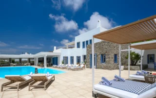 White and stone villa with blue shutters, a turquoise swimming pool and canopy daybeds casting striped shadows across the terrace at Villa Mustique in Mykonos.