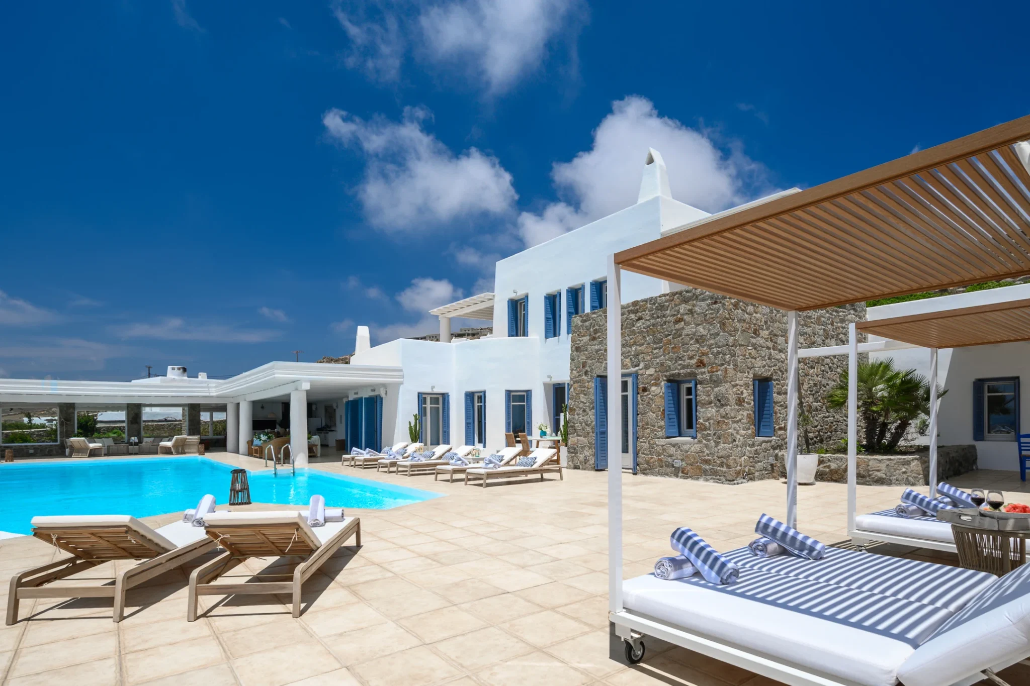 White and stone villa with blue shutters, a turquoise swimming pool and canopy daybeds casting striped shadows across the terrace at Villa Mustique in Mykonos.