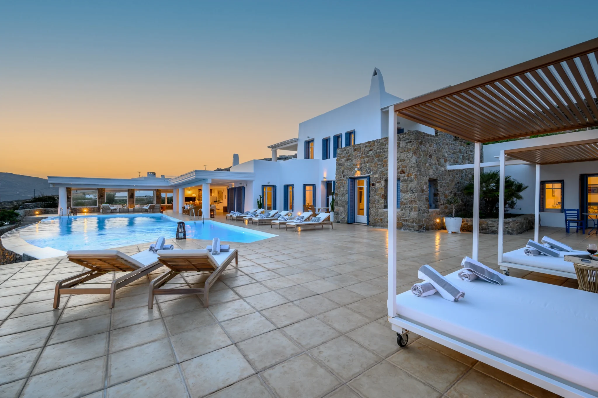White and stone villa with warmly lit windows, a swimming pool and canopy daybeds beneath a sunset sky at Villa Mustique in Mykonos.