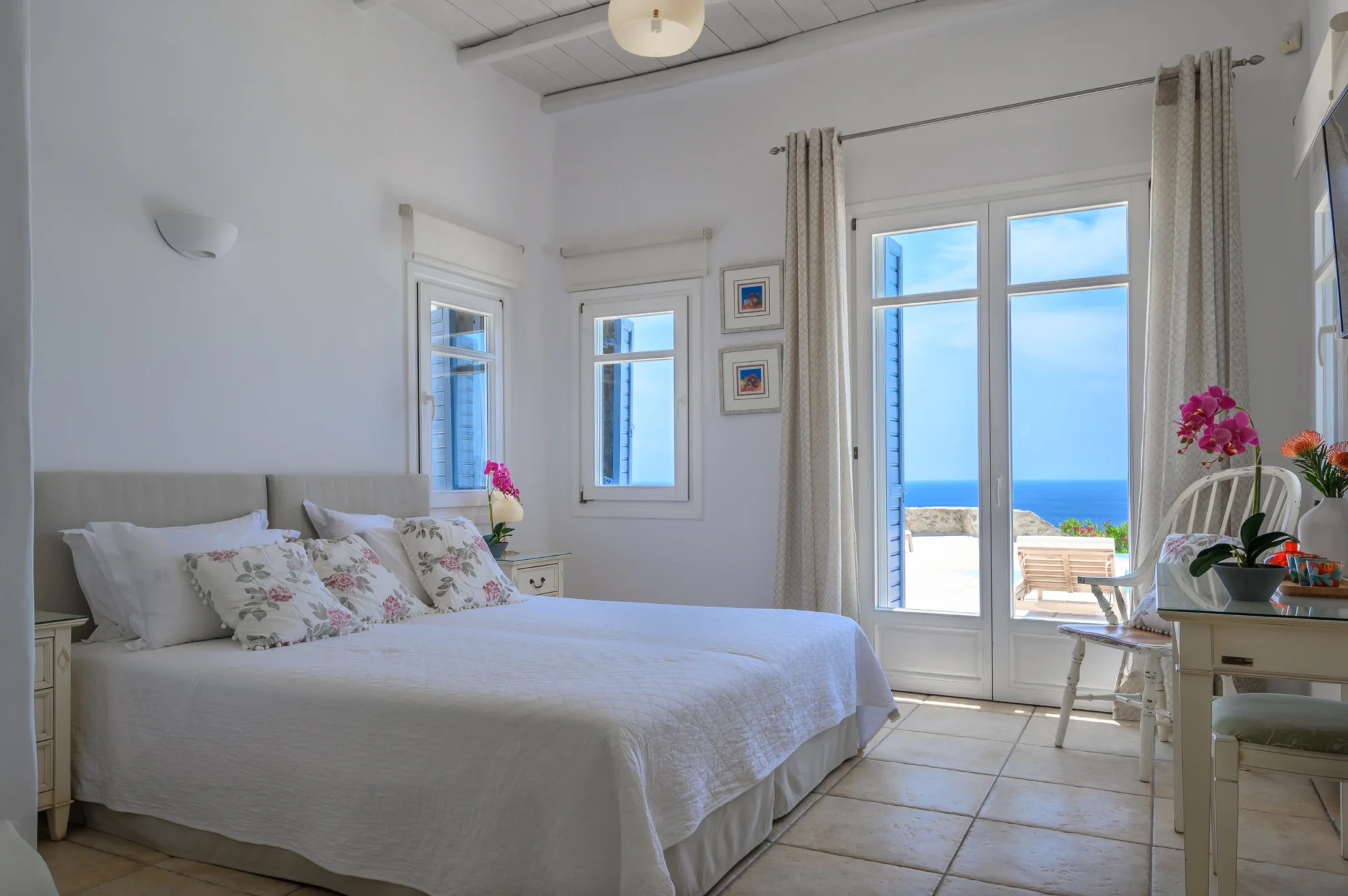 White bedroom with floral cushions and pink orchids, opening through glazed doors to a sea-facing terrace at Villa Mustique in Mykonos.