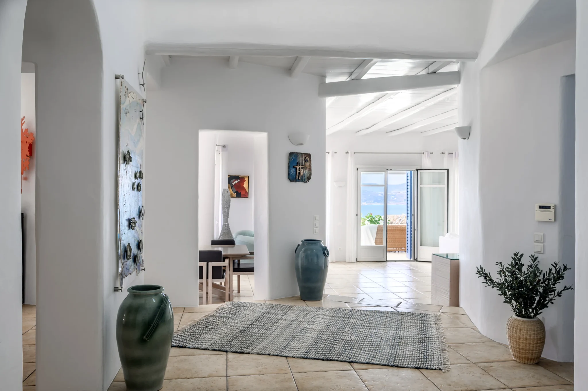 White interior hall with ceramic jars, artwork and a woven rug, looking through the living area towards the terrace at Villa Mustique in Mykonos.