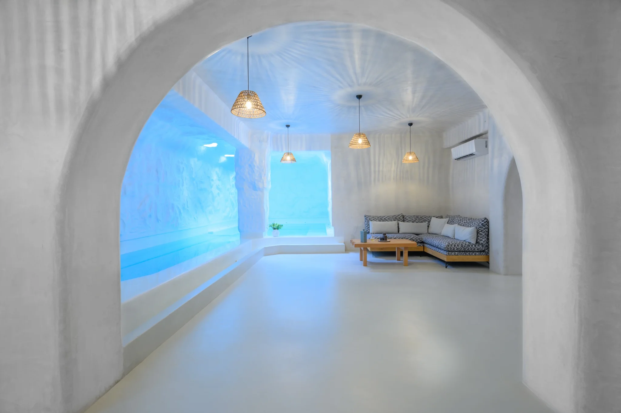 Broad white plaster arch framing a lounge with blue-lit recesses, a patterned sofa and woven pendant lights at Villa Ostraco in Mykonos.