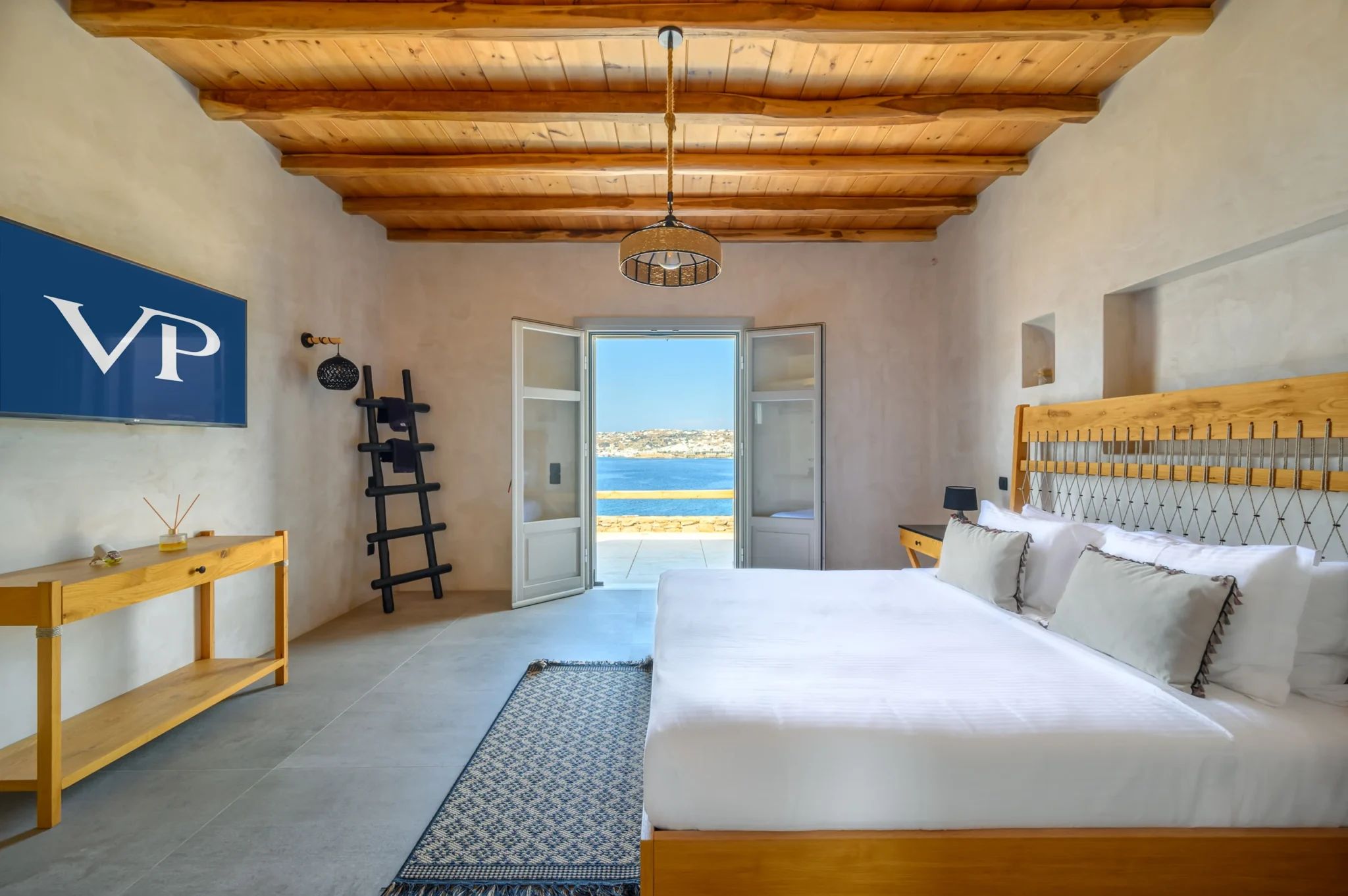 Bedroom with a timber and rope headboard, white bedding and exposed beams, opening through double doors towards the bay at Villa Ostraco in Mykonos.