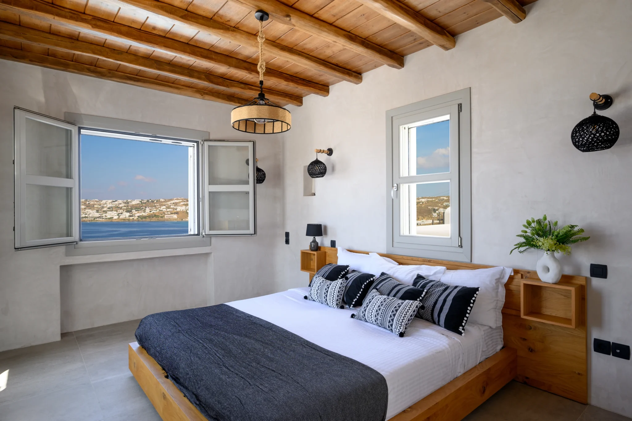 Bedroom with a timber bed, black and white patterned cushions and an open window framing the sea view at Villa Ostraco in Mykonos.