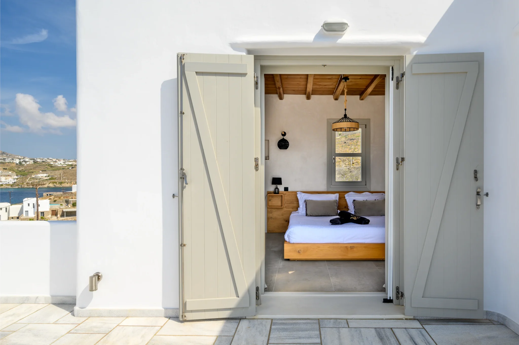 Open pale timber shutters framing a bedroom doorway in the white villa facade, with the bay visible beside the terrace at Villa Ostraco in Mykonos.