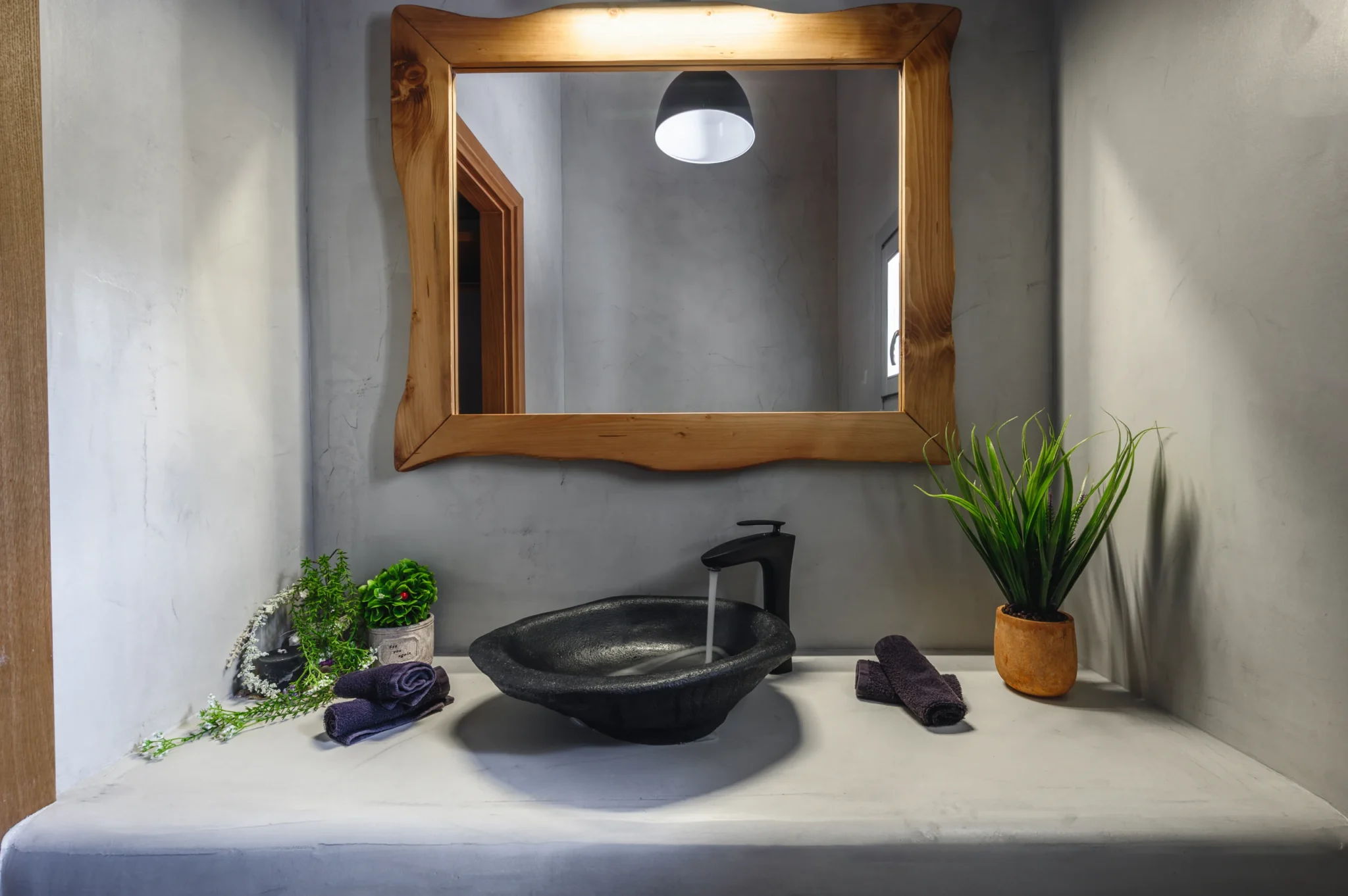 Black vessel basin and tap beneath a timber-framed mirror, with grey plaster walls, plants and rolled towels at Villa Ostraco in Mykonos.