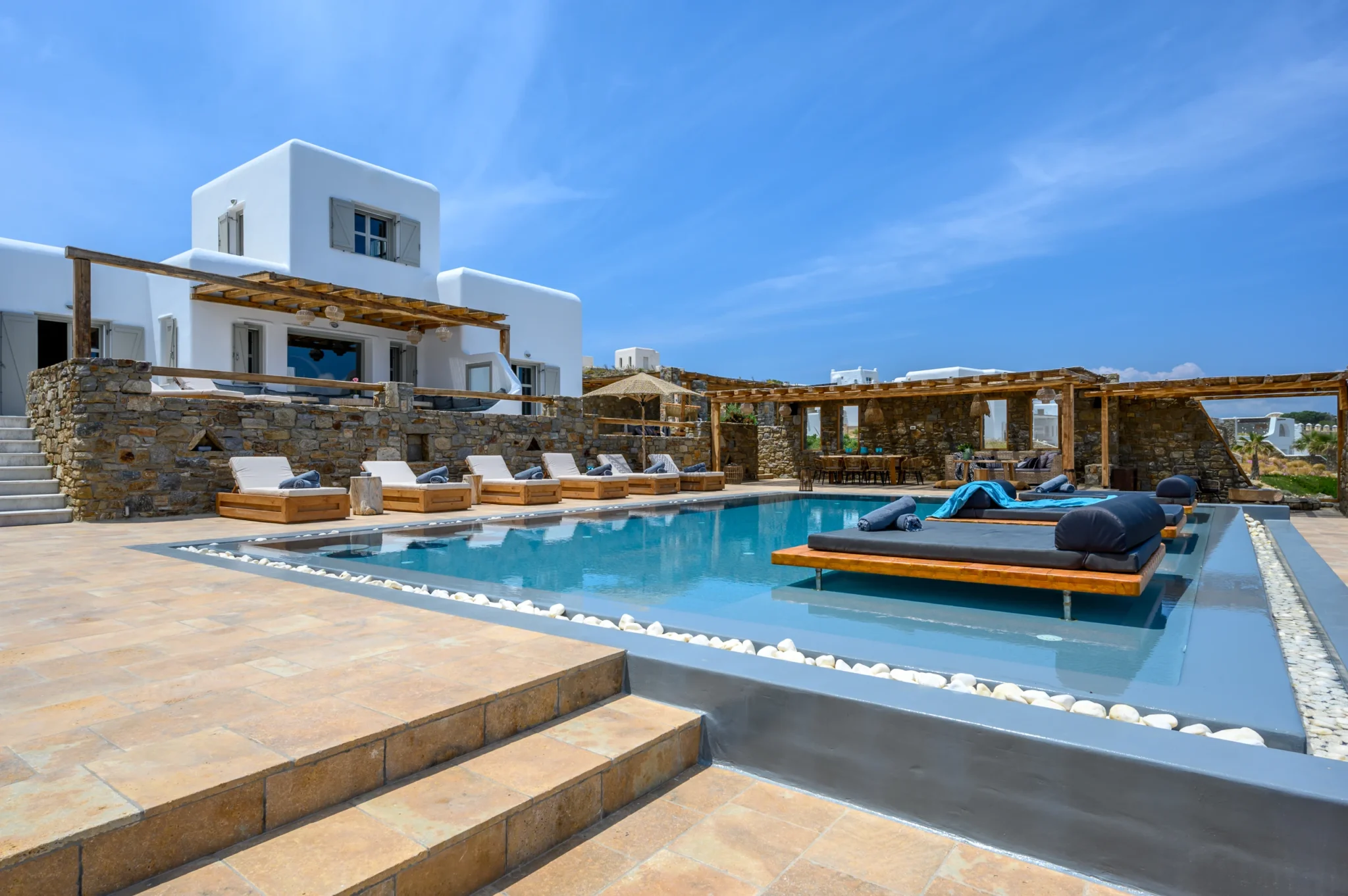 White Cycladic villa above stone terraces, with timber loungers, a swimming pool and daybeds on a shallow water ledge at Villa Ostraco in Mykonos.