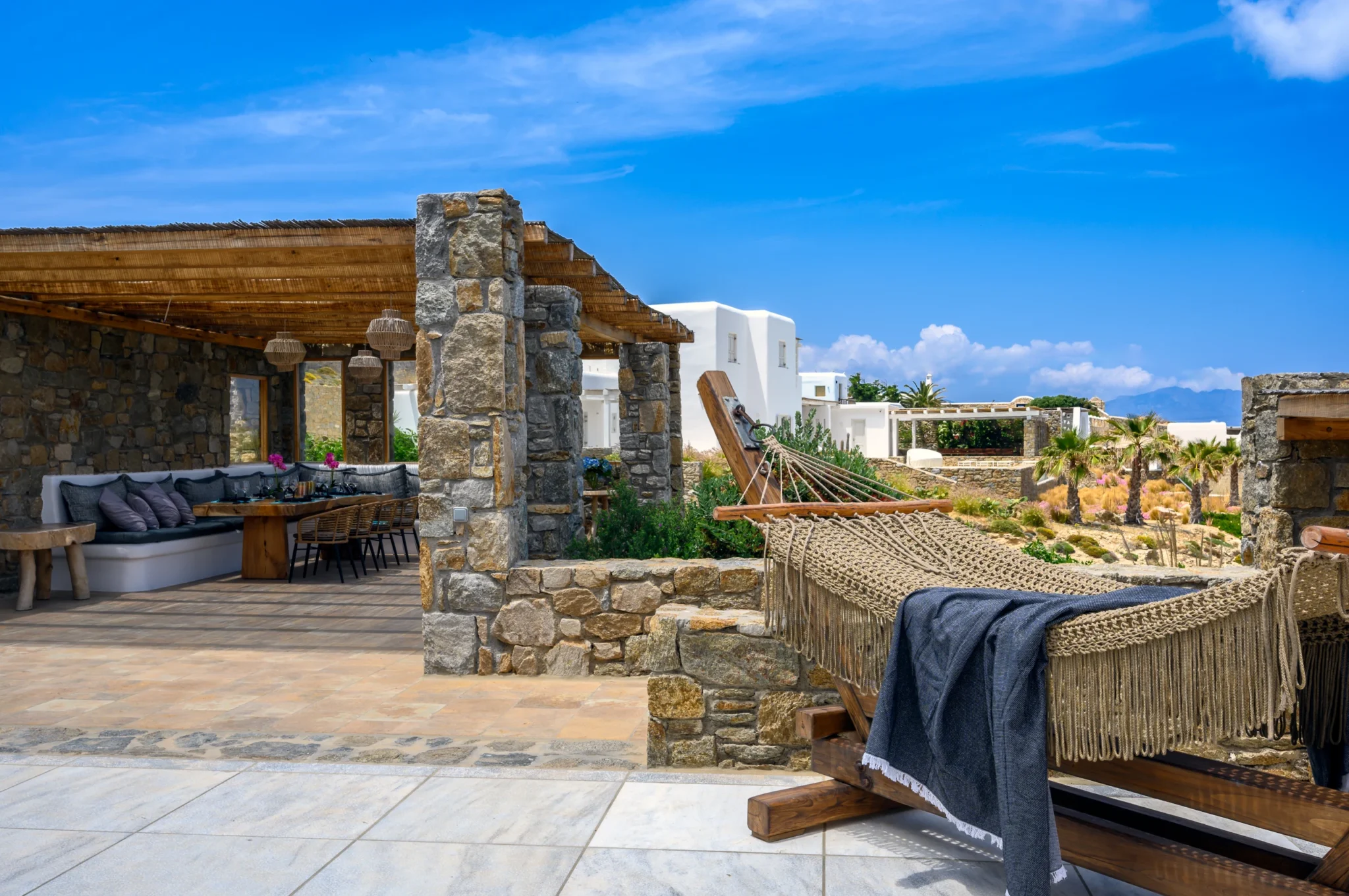 Rope hammock with a blue throw beside a shaded dining terrace with natural stone columns at Villa Ostraco in Mykonos.