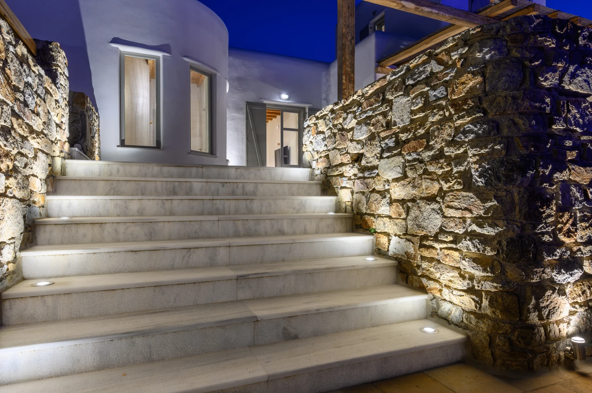 Illuminated white exterior steps between natural stone walls leading to the villa doorway at night at Villa Ostraco in Mykonos.