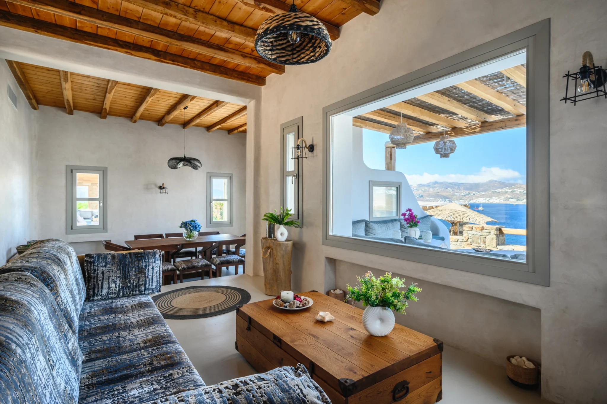 Patterned living room sofa and timber chest table beside a large window framing the shaded terrace and sea at Villa Ostraco in Mykonos.