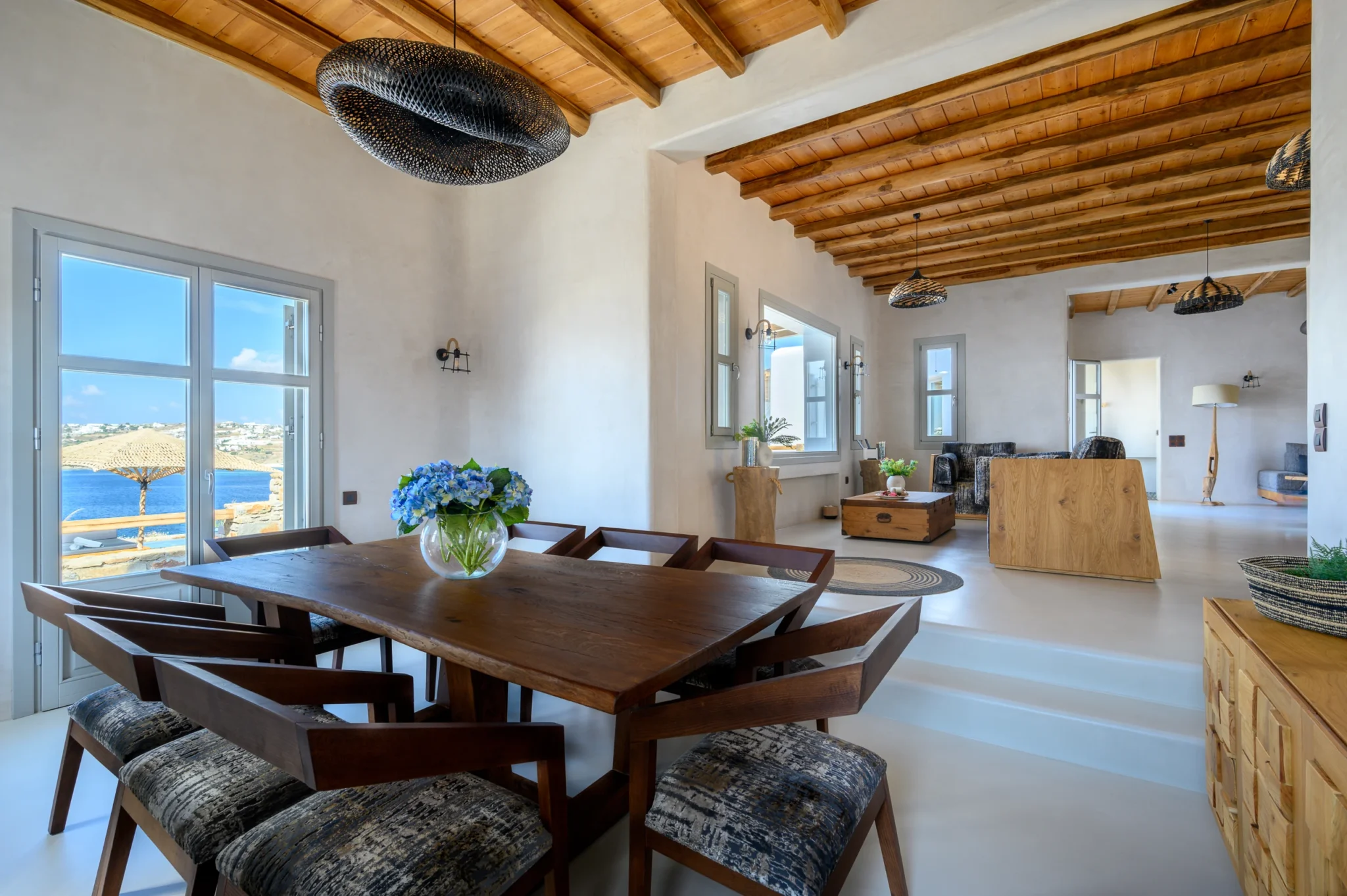 Timber dining table with blue hydrangeas leading into a living room beneath exposed timber beams and woven pendant lights at Villa Ostraco in Mykonos.