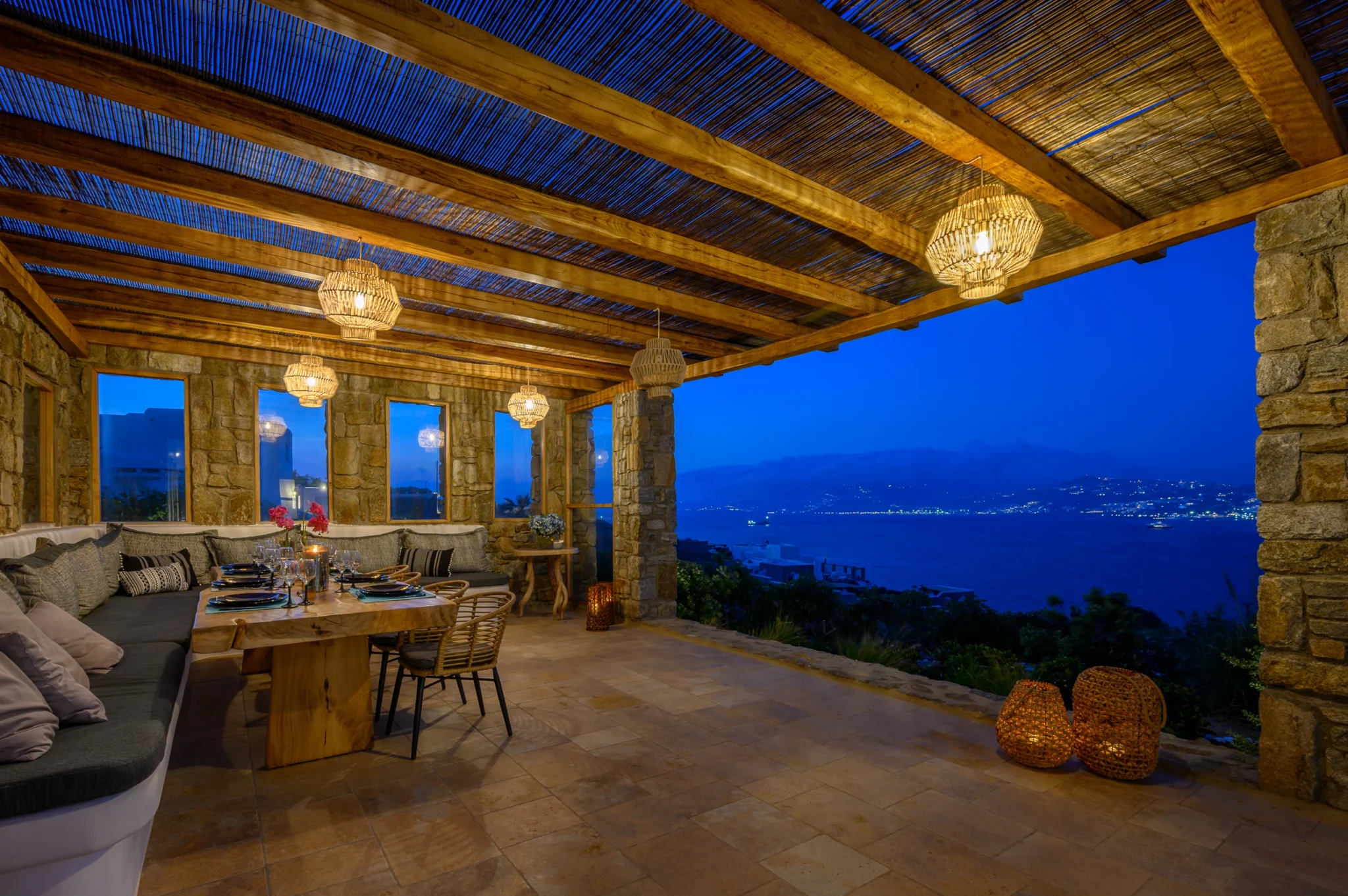 Set outdoor dining table and cushioned bench beneath warm woven pendant lights, overlooking Mykonos bay at blue hour at Villa Ostraco in Mykonos.