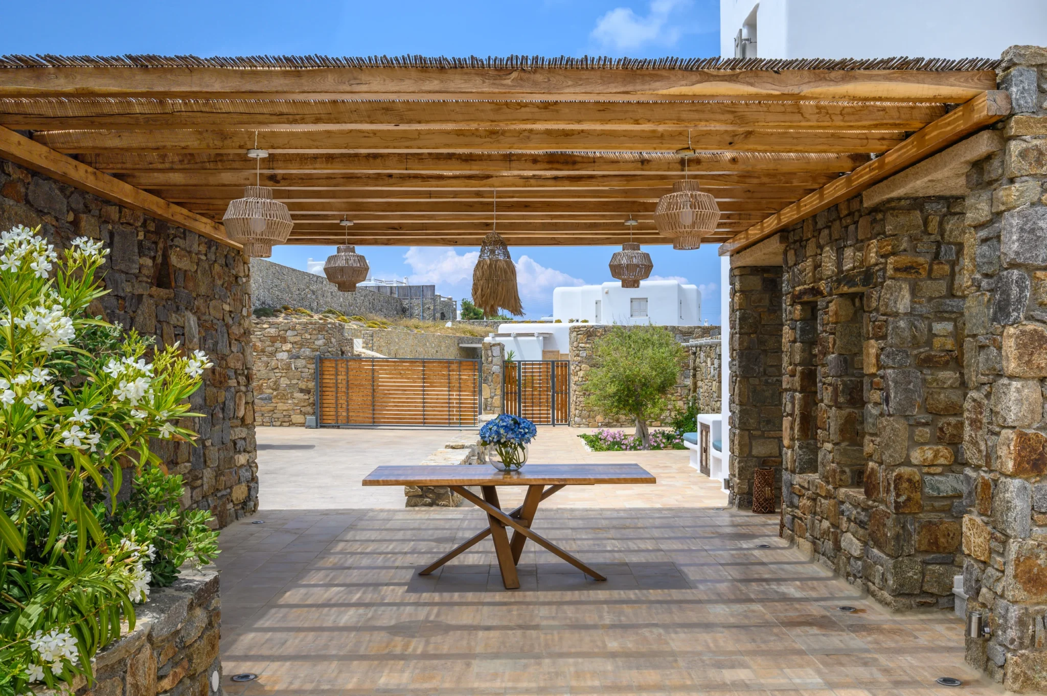 Stone courtyard beneath a timber and reed pergola, with a flower-topped table, planting and timber entrance gates at Villa Ostraco in Mykonos.