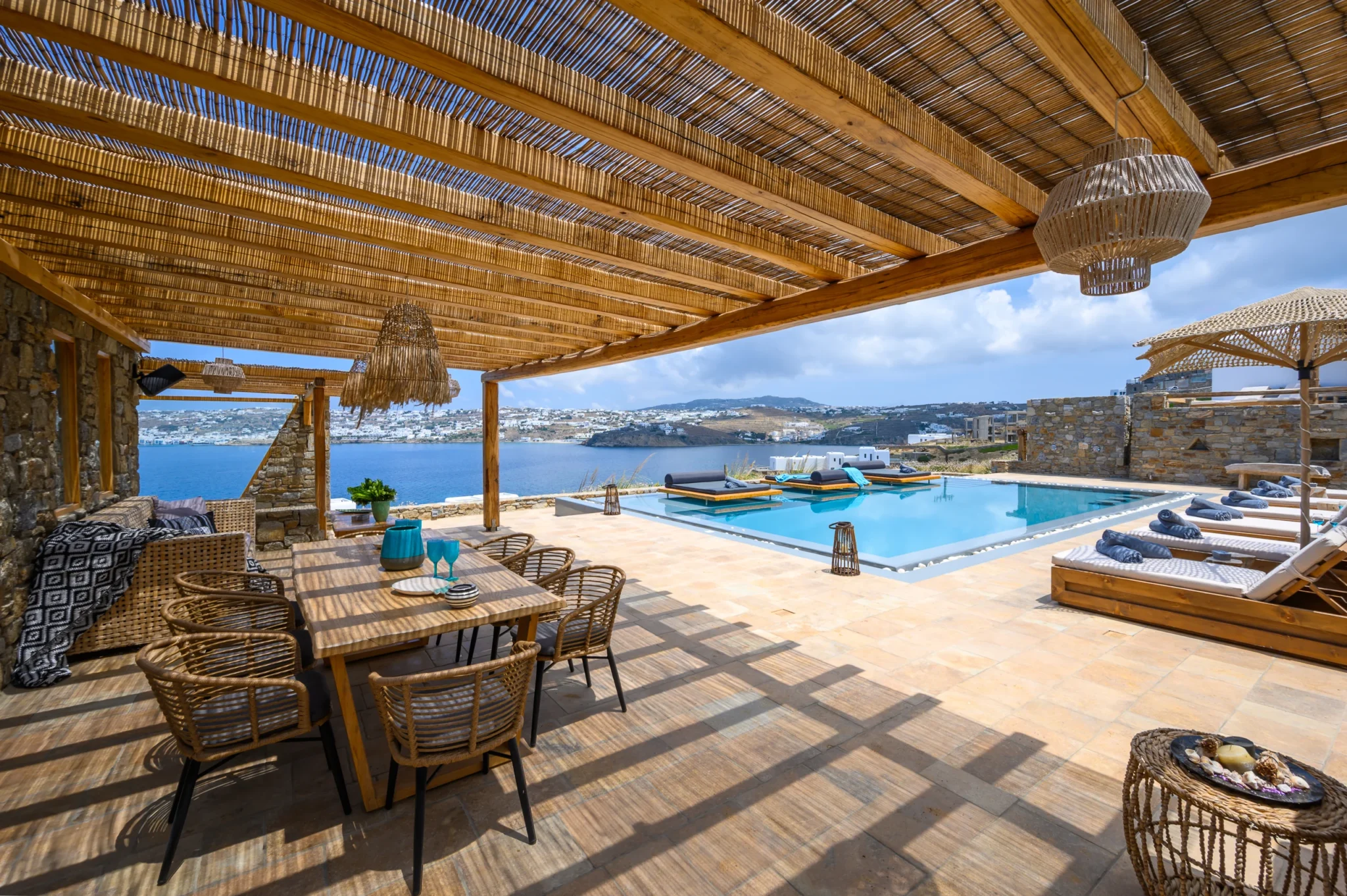 Timber dining table and woven chairs beneath a reed pergola, looking across the pool and loungers towards the bay at Villa Ostraco in Mykonos.