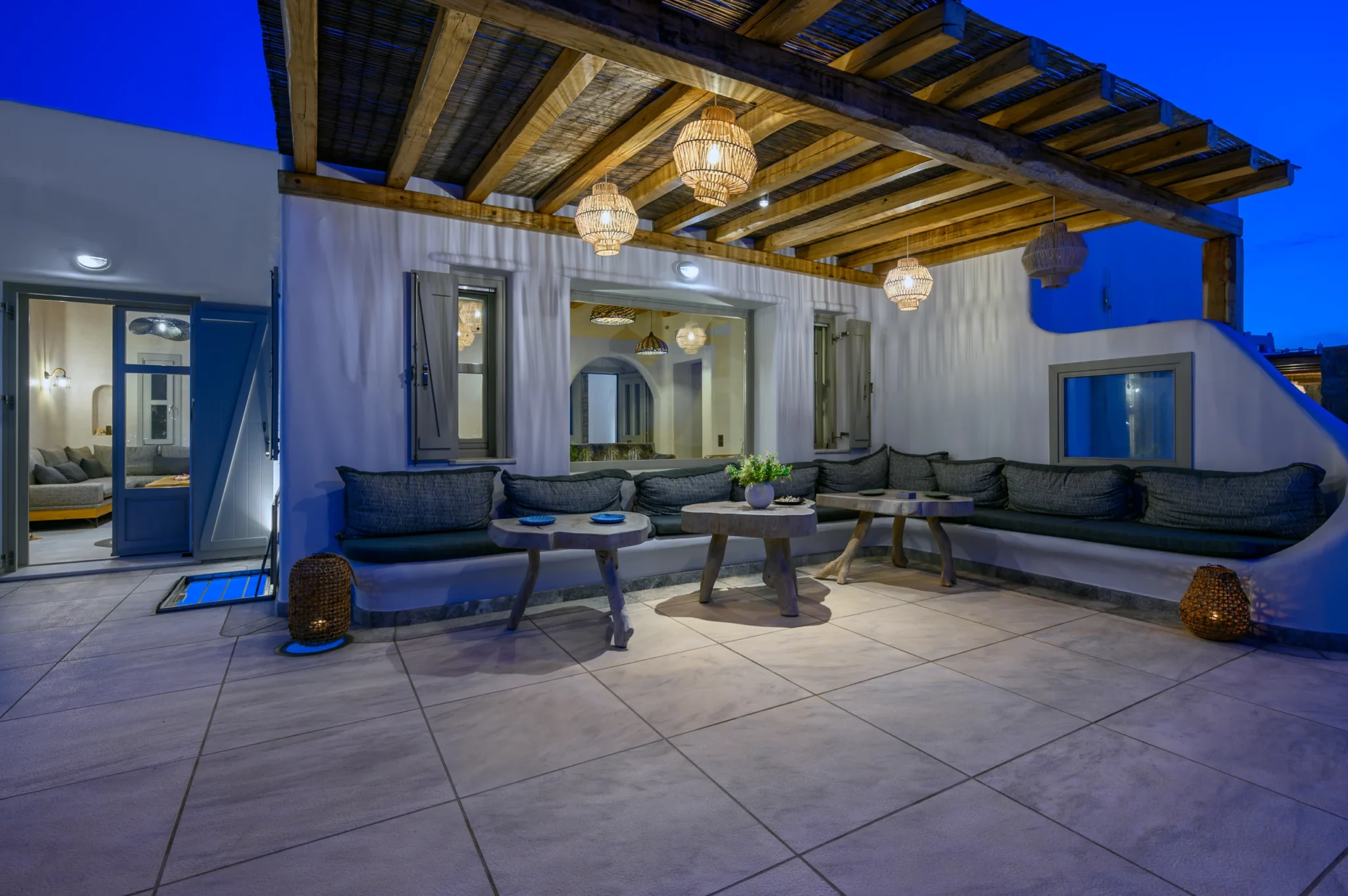 Built-in cushioned terrace seating and timber tables beneath illuminated woven pendant lights at blue hour at Villa Ostraco in Mykonos.