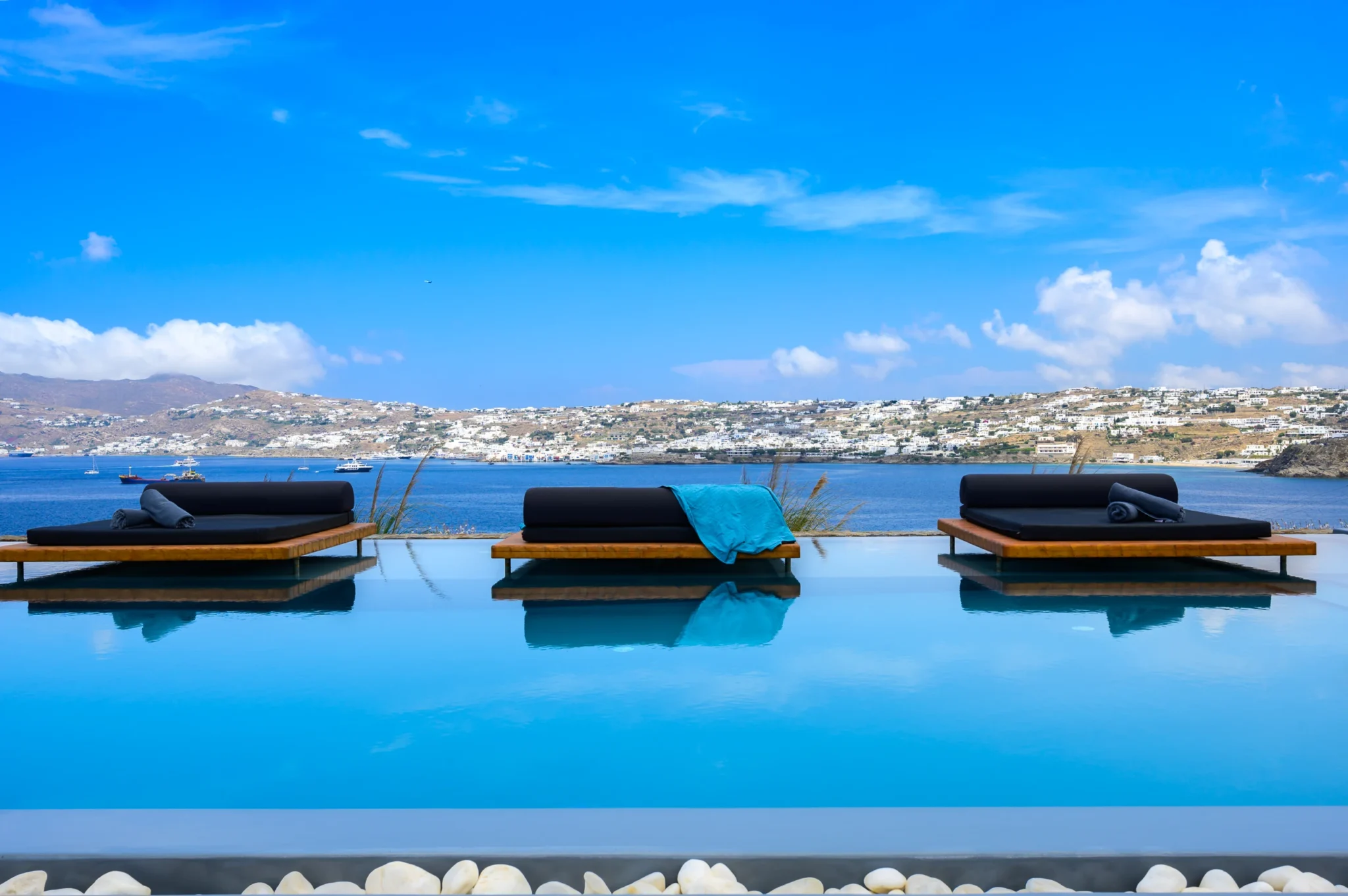 Three cushioned timber daybeds reflected in the swimming pool, with the sea and Mykonos coastline beyond at Villa Ostraco in Mykonos.