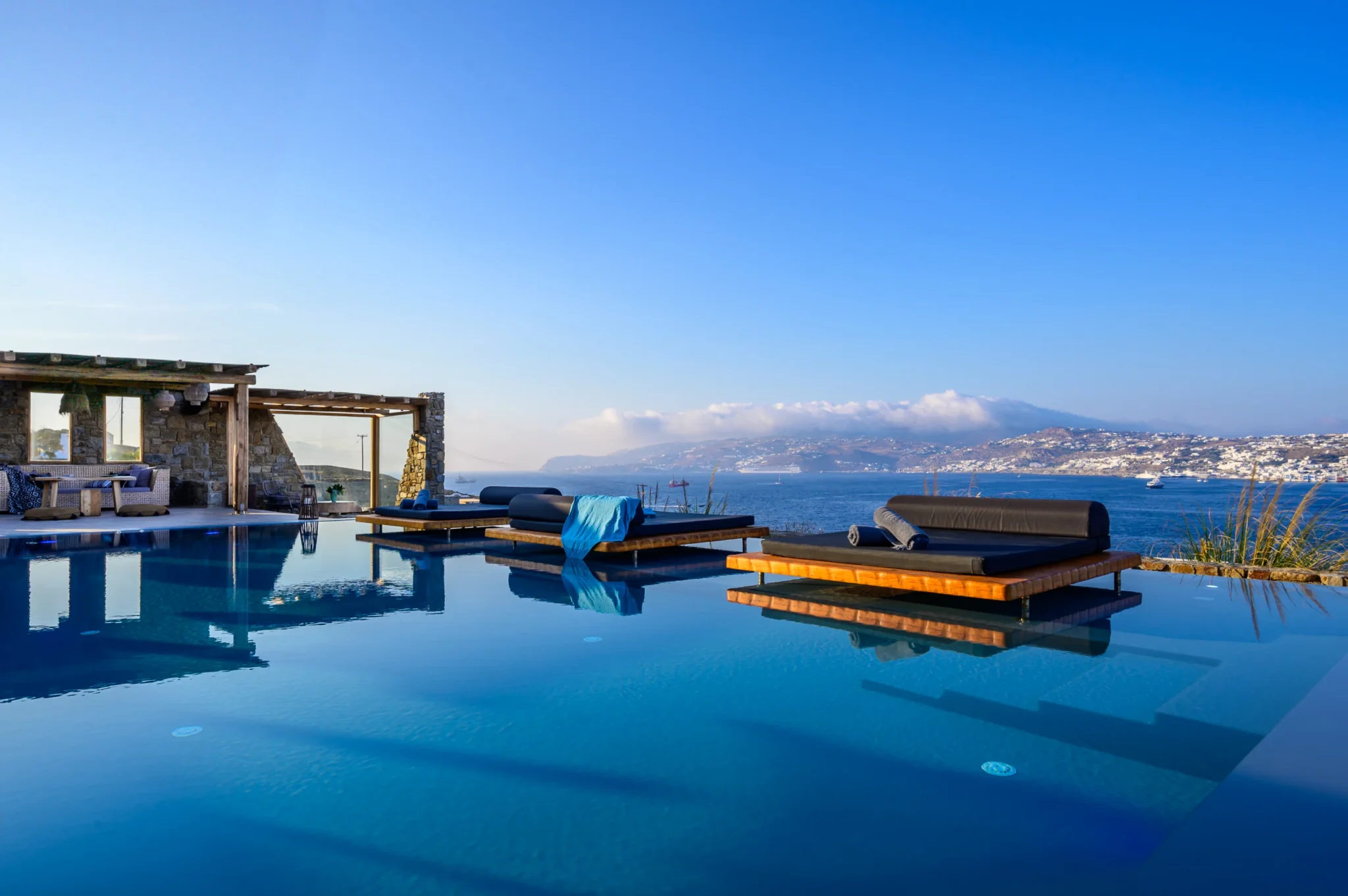 Timber platform daybeds reflected in still pool water beside a stone pavilion, with the bay in soft evening light at Villa Ostraco in Mykonos.