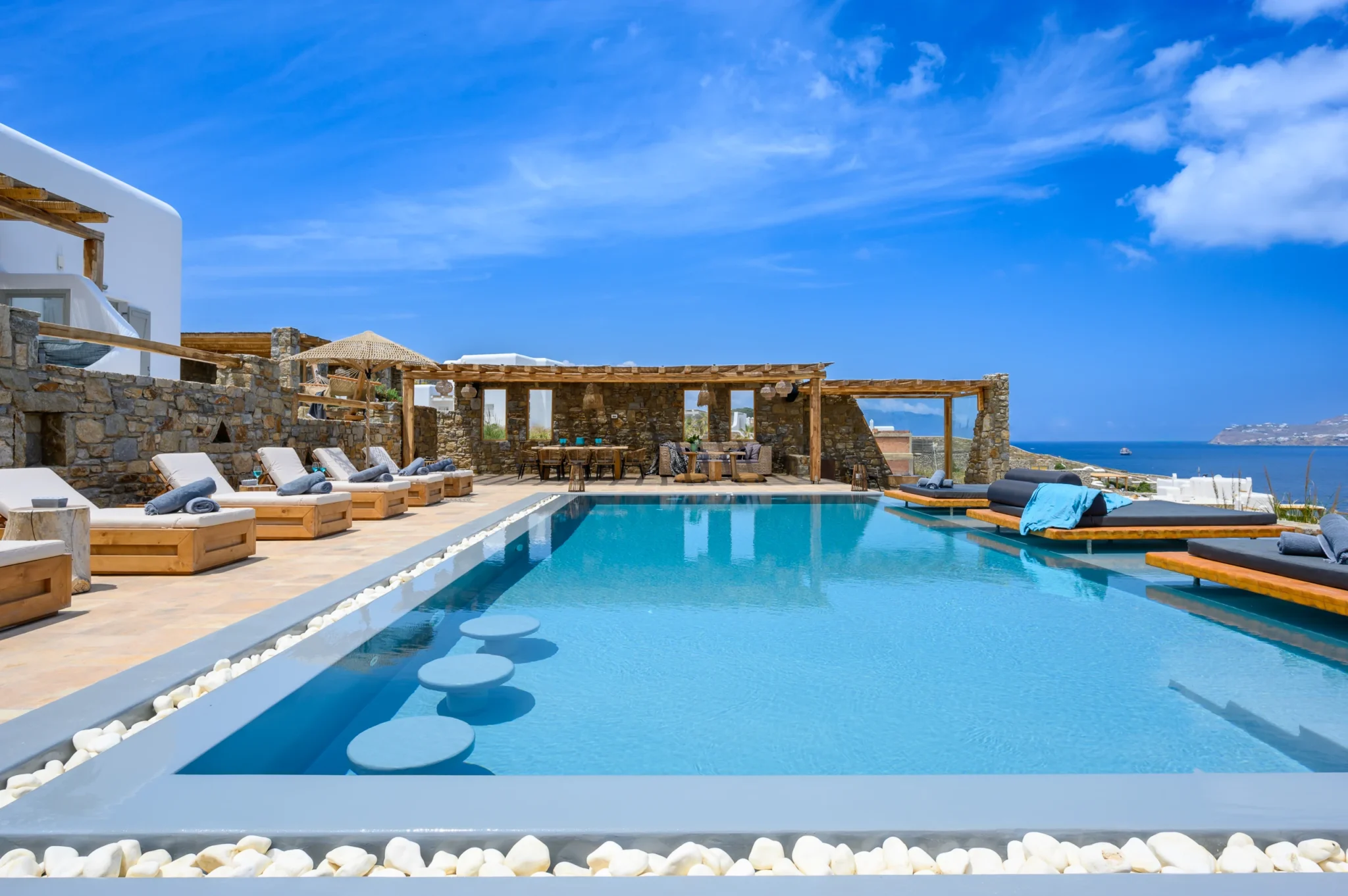 Swimming pool with round submerged stools, timber platform daybeds and a stone dining pavilion beside the sea view at Villa Ostraco in Mykonos.