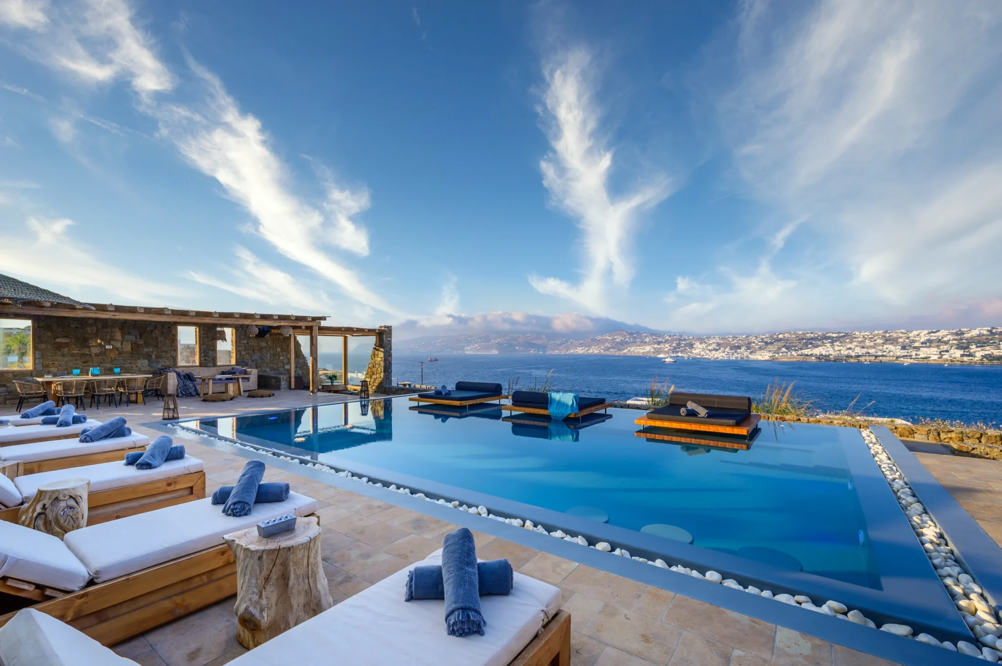 White-cushioned timber loungers beside the swimming pool and platform daybeds, overlooking the bay in evening light at Villa Ostraco in Mykonos.