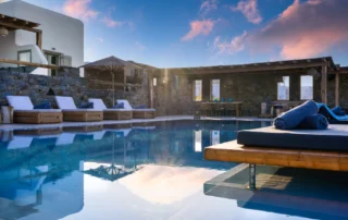 Swimming pool reflecting pink sunset clouds, with a cushioned timber daybed, sun loungers and a stone dining pergola at Villa Ostraco in Mykonos.