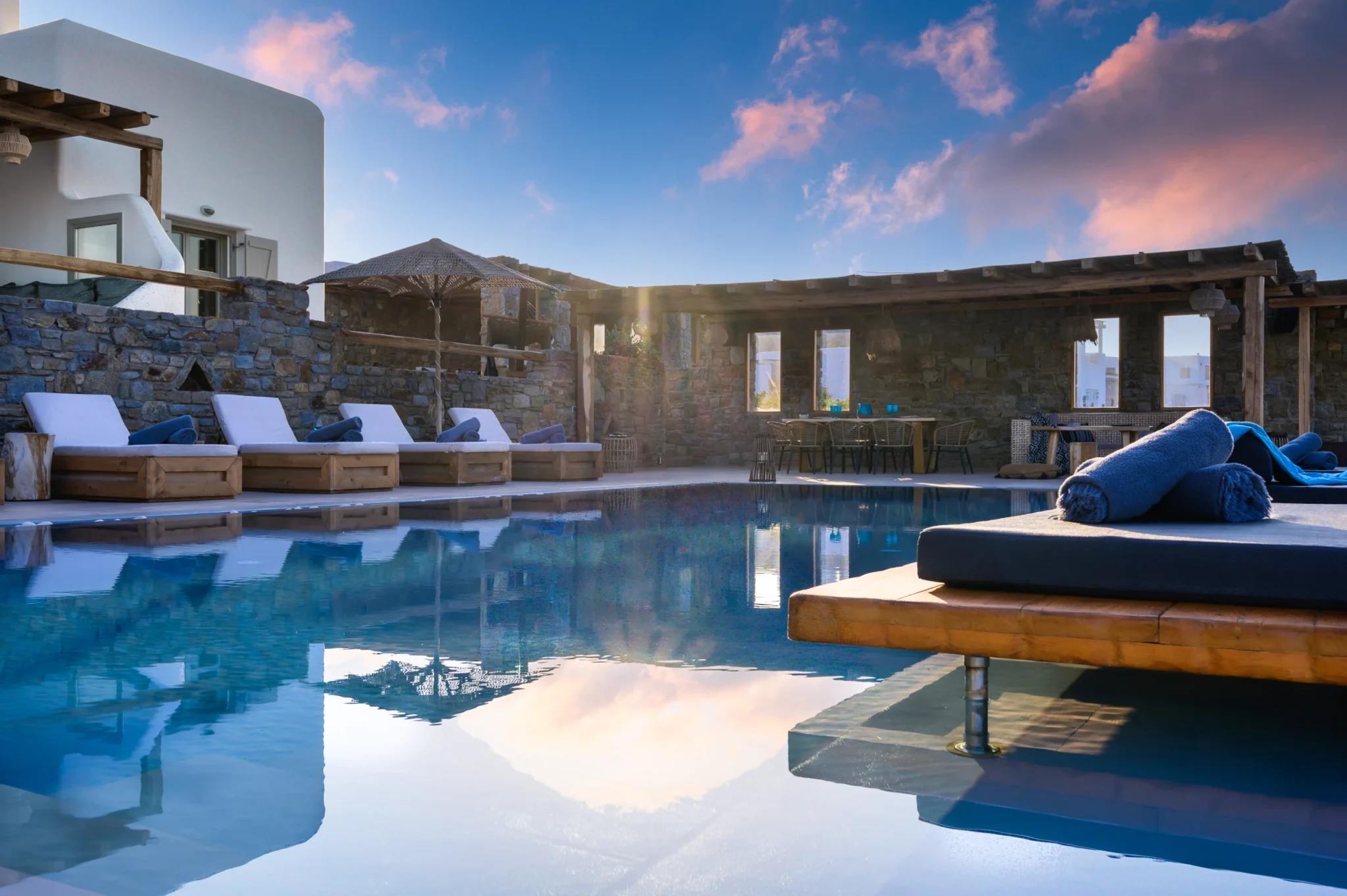 Swimming pool reflecting pink sunset clouds, with a cushioned timber daybed, sun loungers and a stone dining pergola at Villa Ostraco in Mykonos.