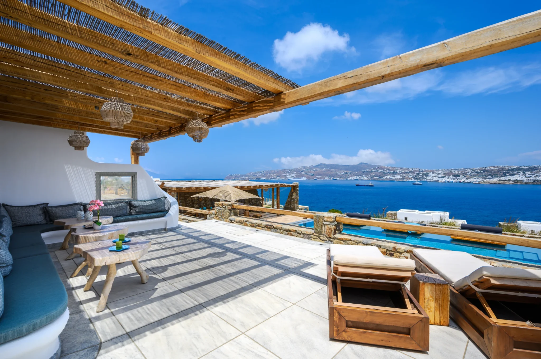 Built-in cushioned seating and timber tables beneath a reed pergola, overlooking the pool and Mykonos bay at Villa Ostraco in Mykonos.
