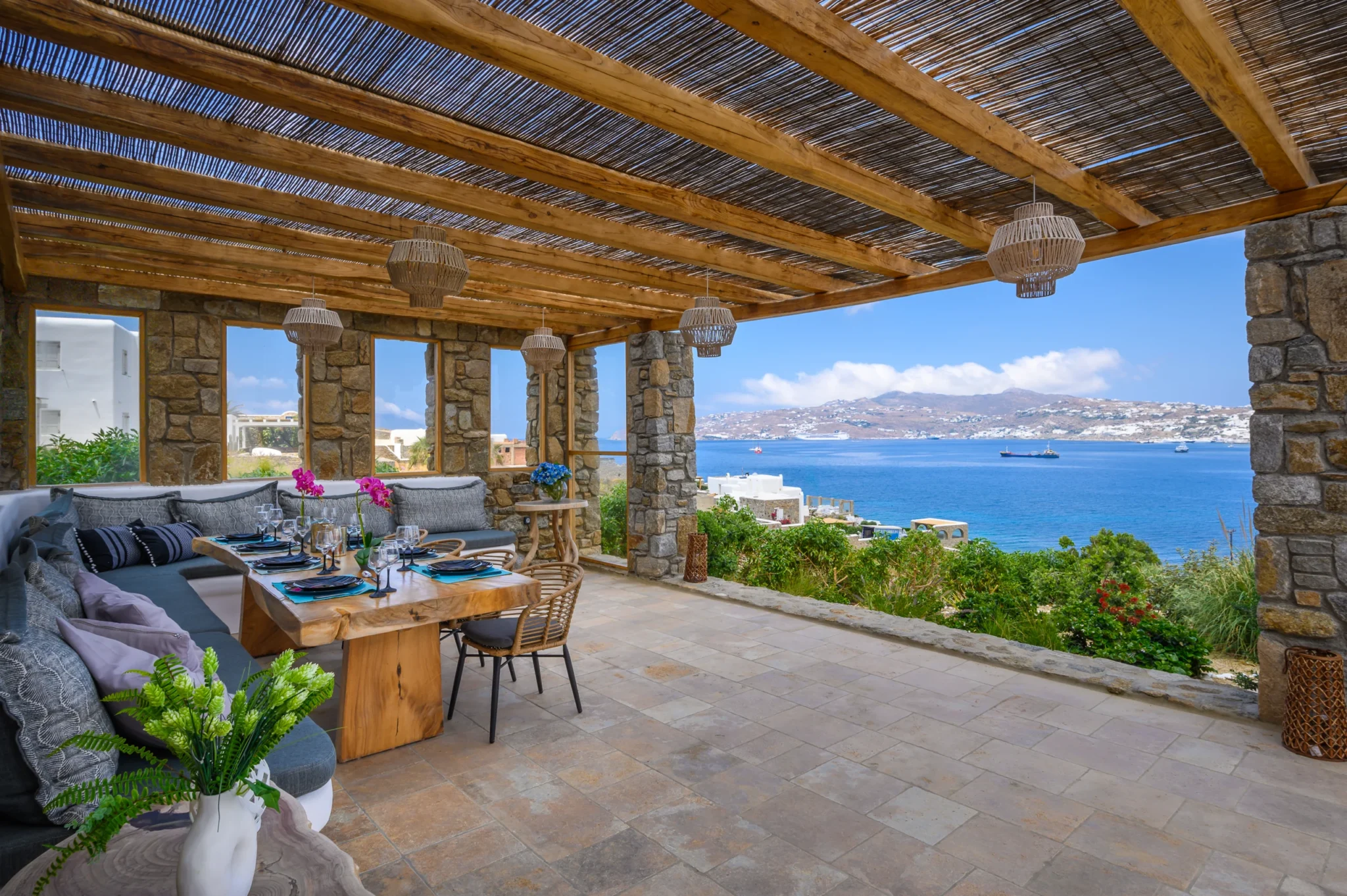Set timber dining table and cushioned built-in seating beneath a stone and timber pavilion overlooking the sea at Villa Ostraco in Mykonos.