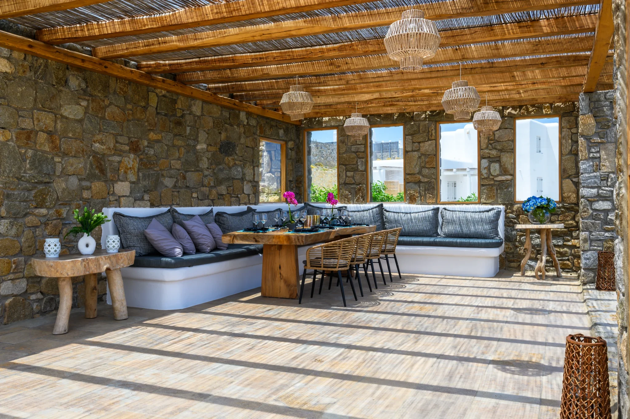 Dining table with woven chairs and wraparound cushioned seating beneath woven pendant lights and a reed pergola casting striped shadows at Villa Ostraco in Mykonos.