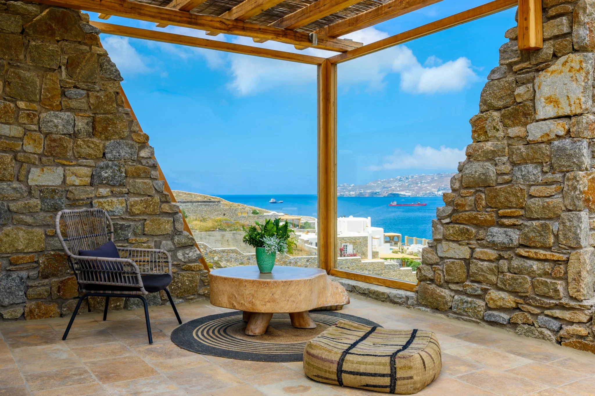 Woven armchair, timber table and low ottoman beside a glass corner screen framing the sea between stone walls at Villa Ostraco in Mykonos.