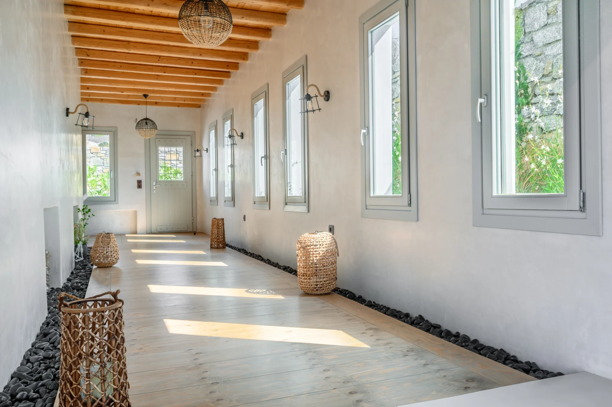 White corridor with grey-framed windows, a timber floor edged with black pebbles and woven lanterns in patches of sunlight at Villa Ostraco in Mykonos.