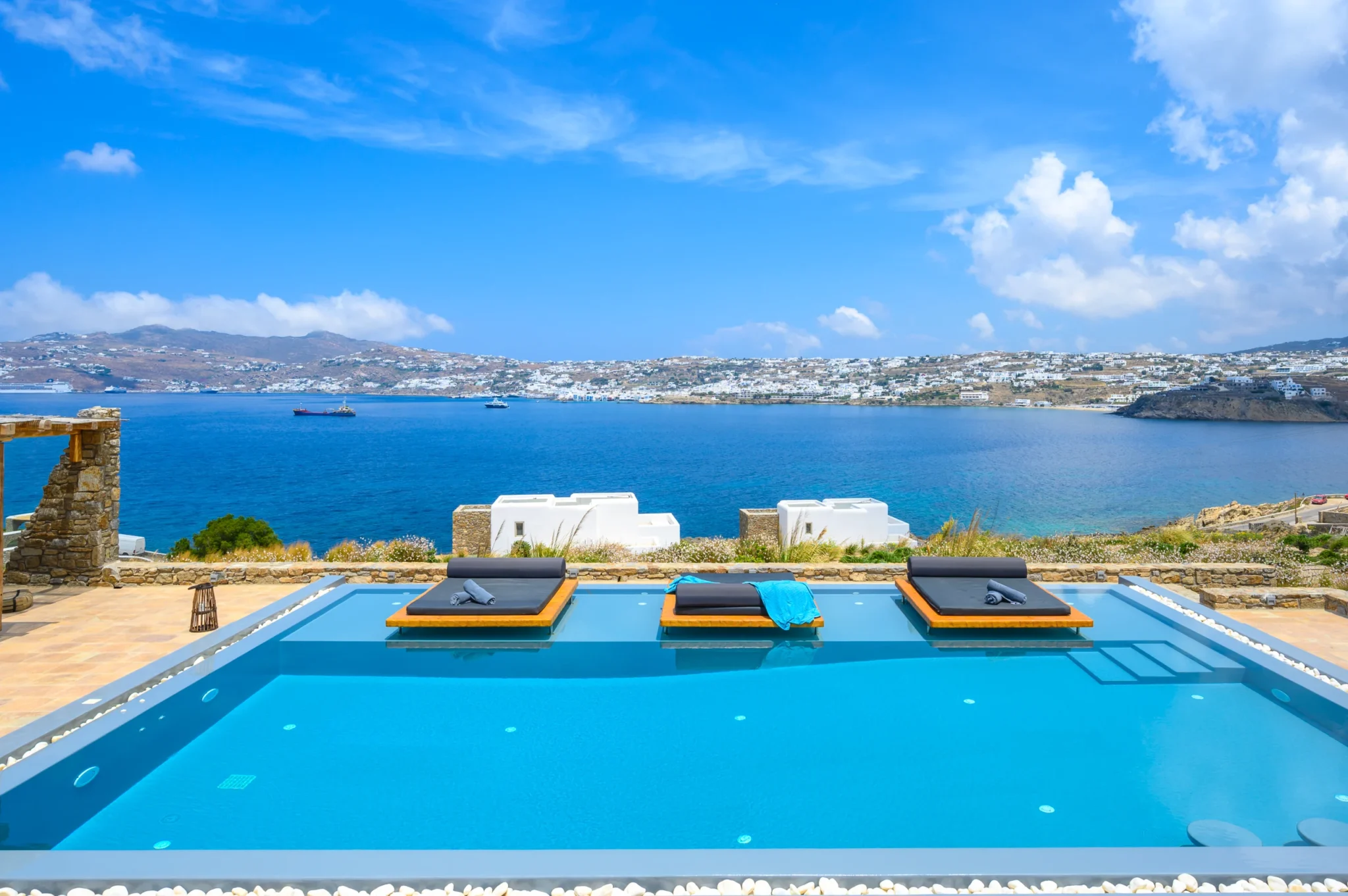 Swimming pool and three timber platform daybeds overlooking the sea and the white buildings across Mykonos bay at Villa Ostraco in Mykonos.