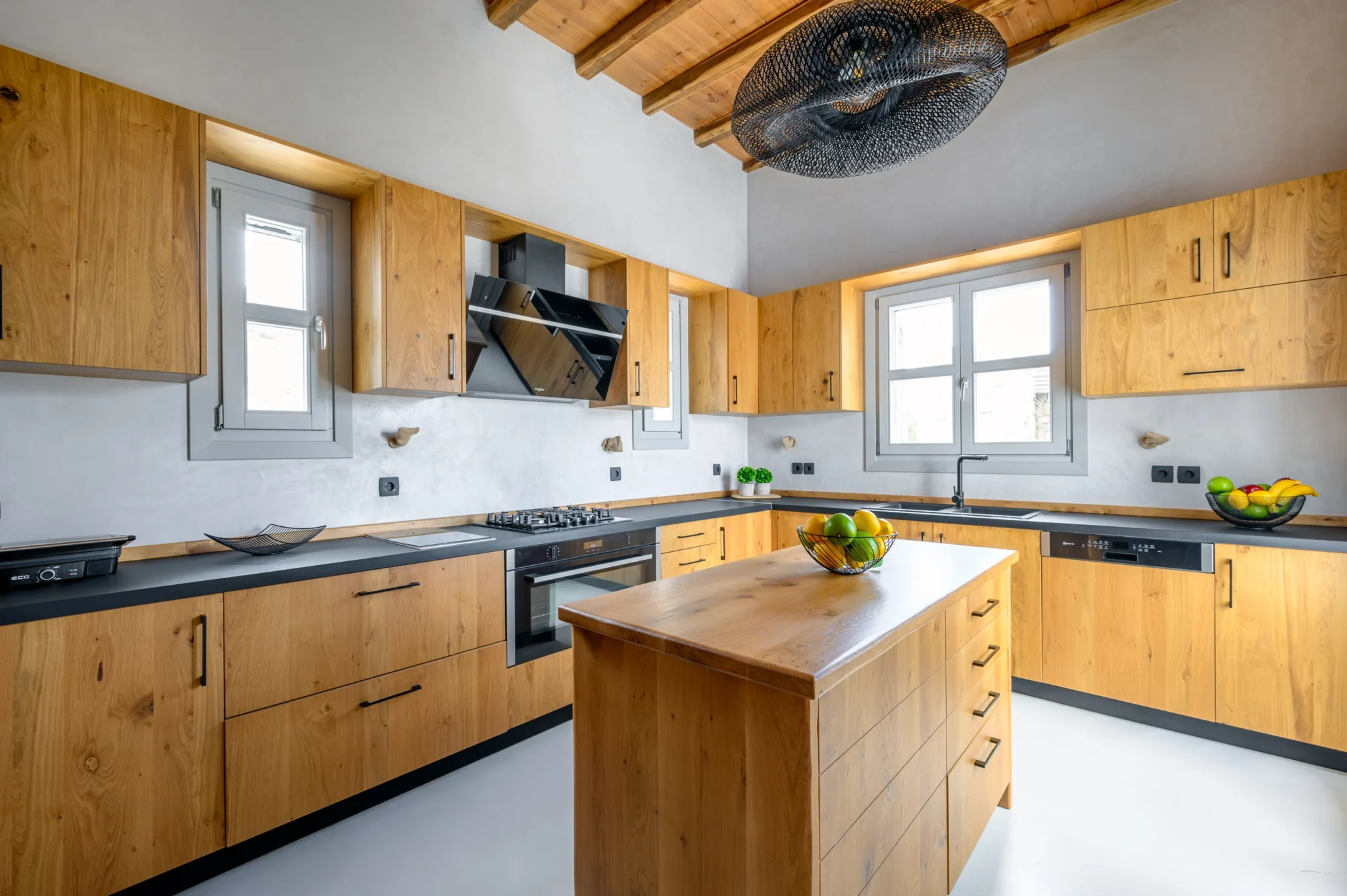 Kitchen with natural timber cabinetry and an island, dark worktops, grey-framed windows and exposed ceiling beams at Villa Ostraco in Mykonos.